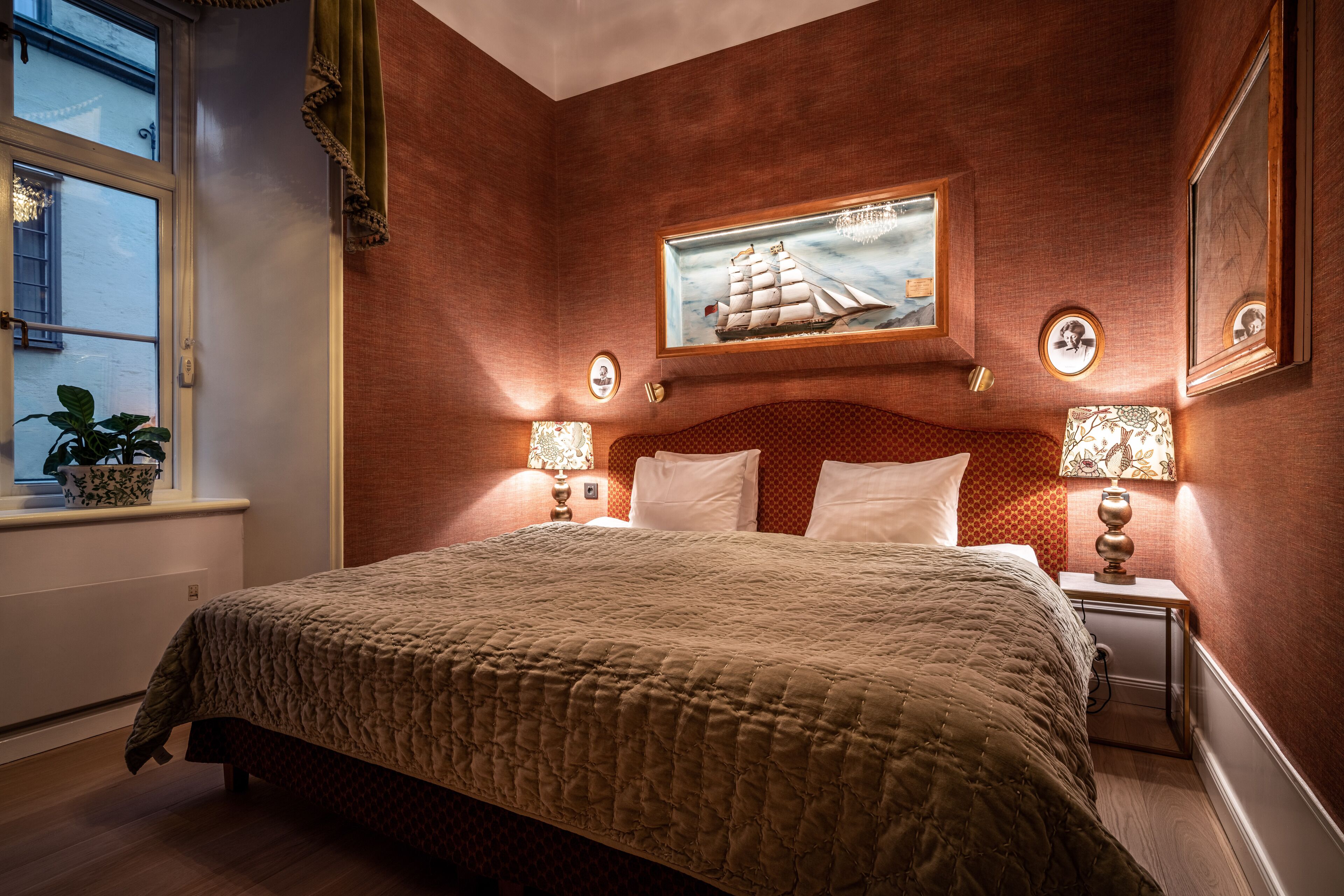 Ein Schlafzimmer mit einem großen Bett, zwei Nachttischen mit Lampen, einem Fenster mit einer Pflanze und einem Wandbild eines Schiffes.