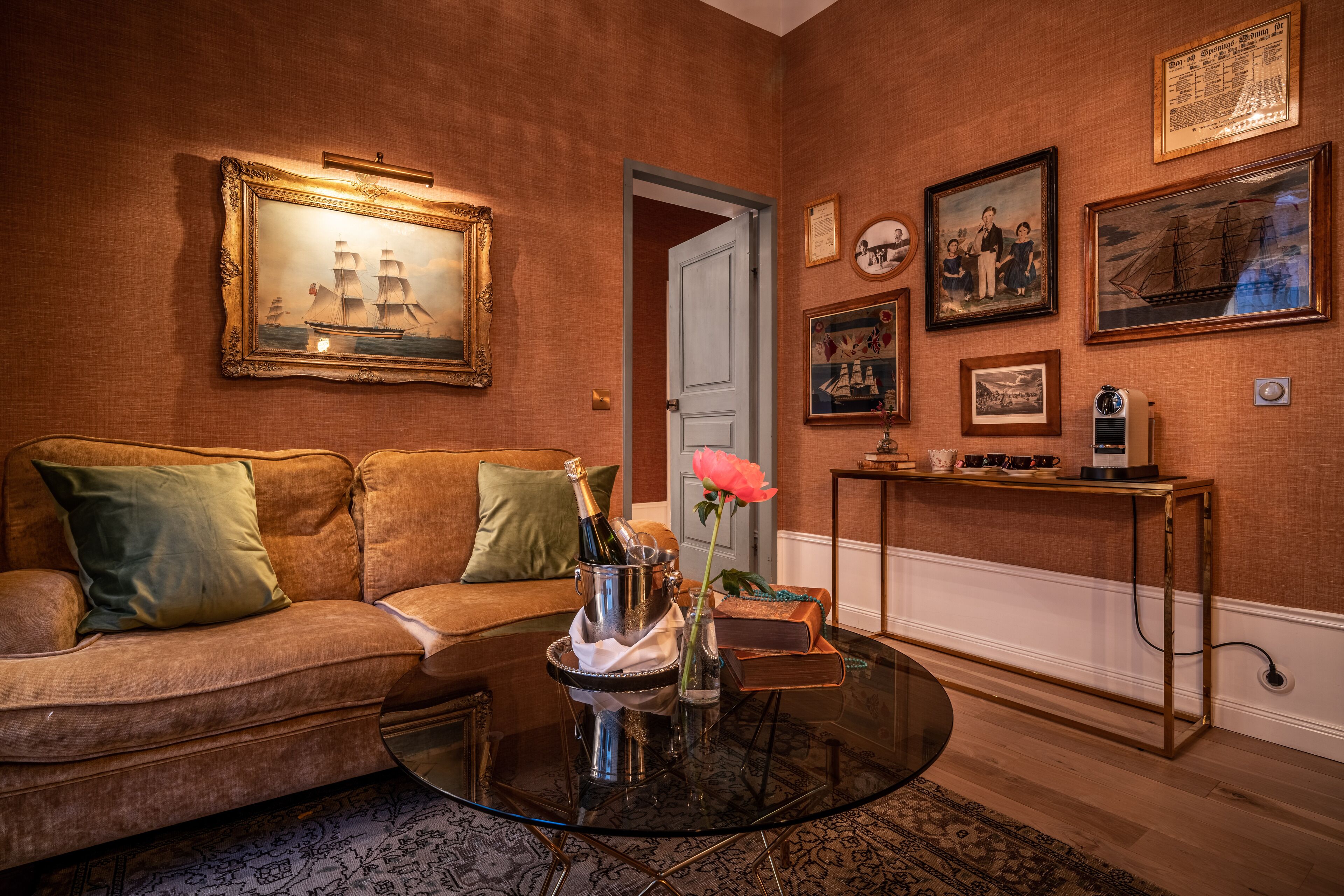 A cozy living room with a sofa, a glass coffee table with a champagne bottle and flowers, and a wall adorned with framed artwork.