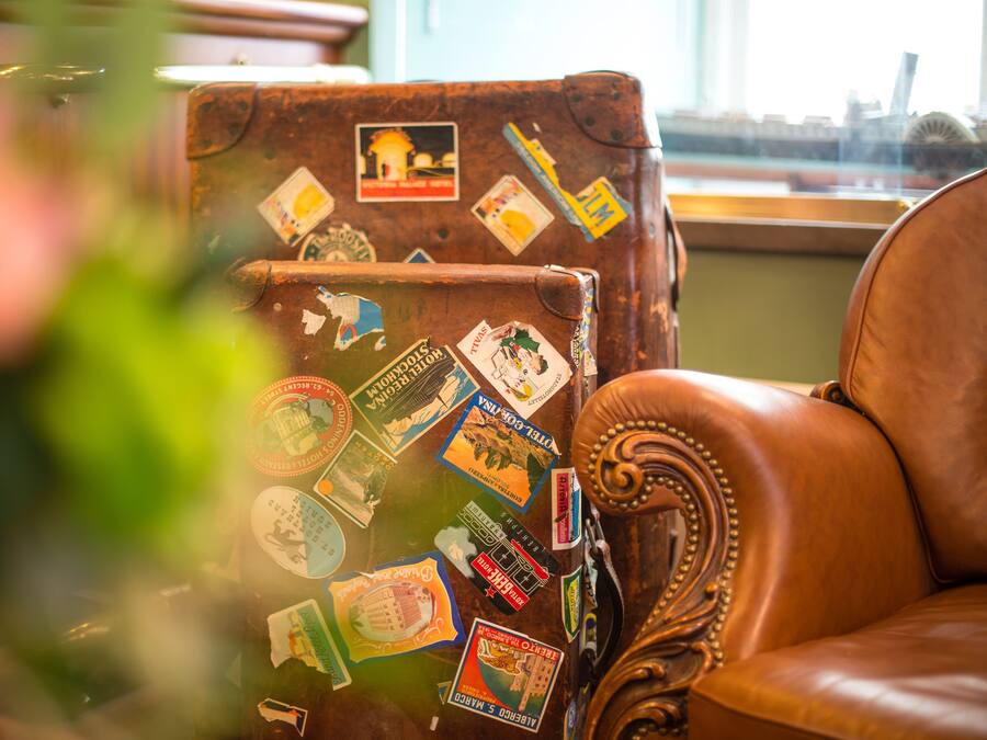 Una maleta antigua con etiquetas de viaje, un sillón de cuero y una ventana con vistas.