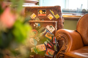 Una maleta antigua con etiquetas de viaje, un sillón de cuero y una ventana con vistas.