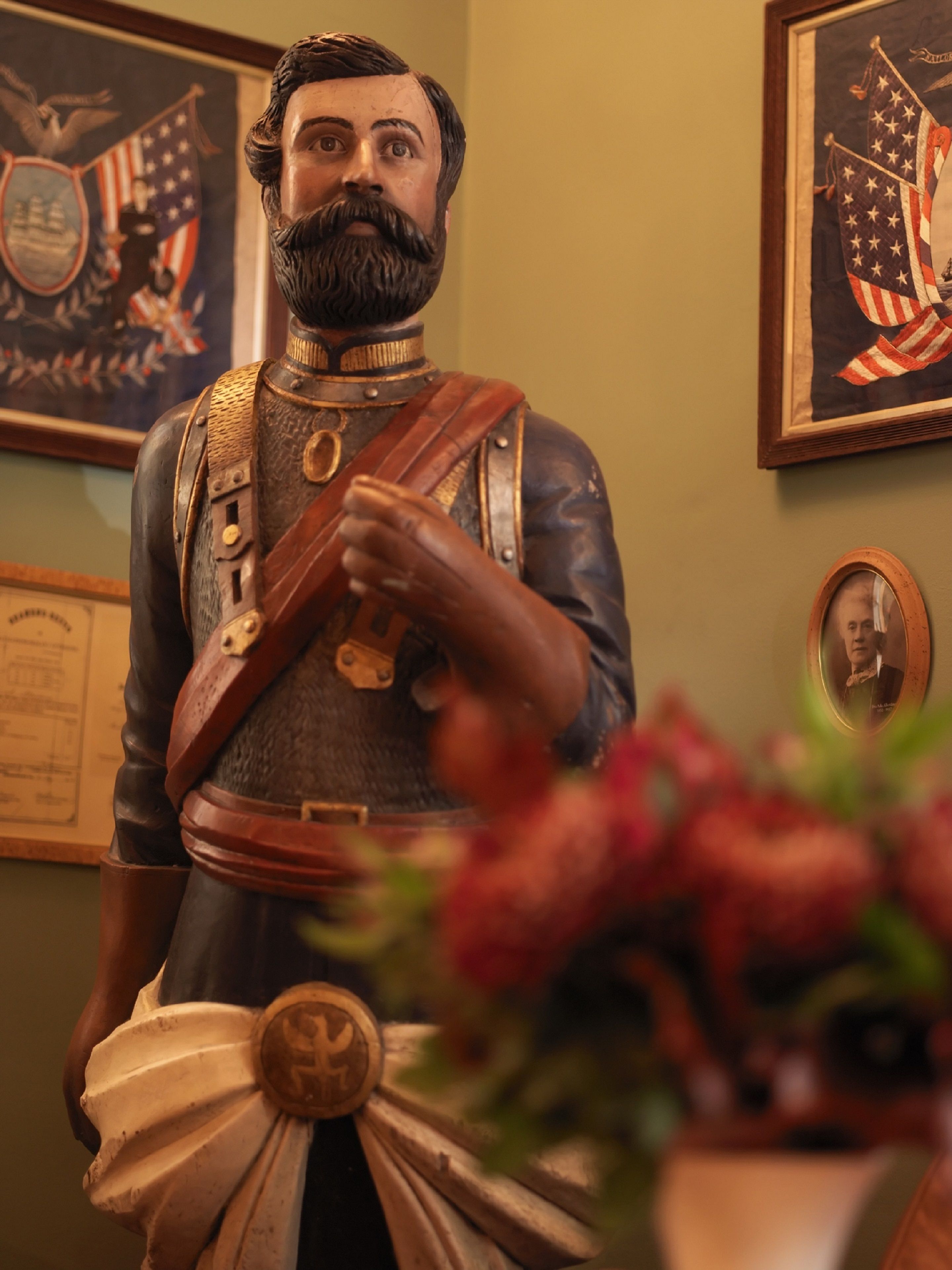 Estatua de una figura histórica con uniforme militar, medallas y espada, frente a una pared con cuadros enmarcados y una placa conmemorativa.