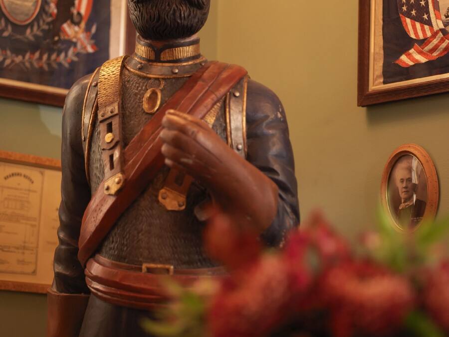 Estatua de una figura histórica con uniforme militar, medallas y espada, frente a una pared con cuadros enmarcados y una placa conmemorativa.