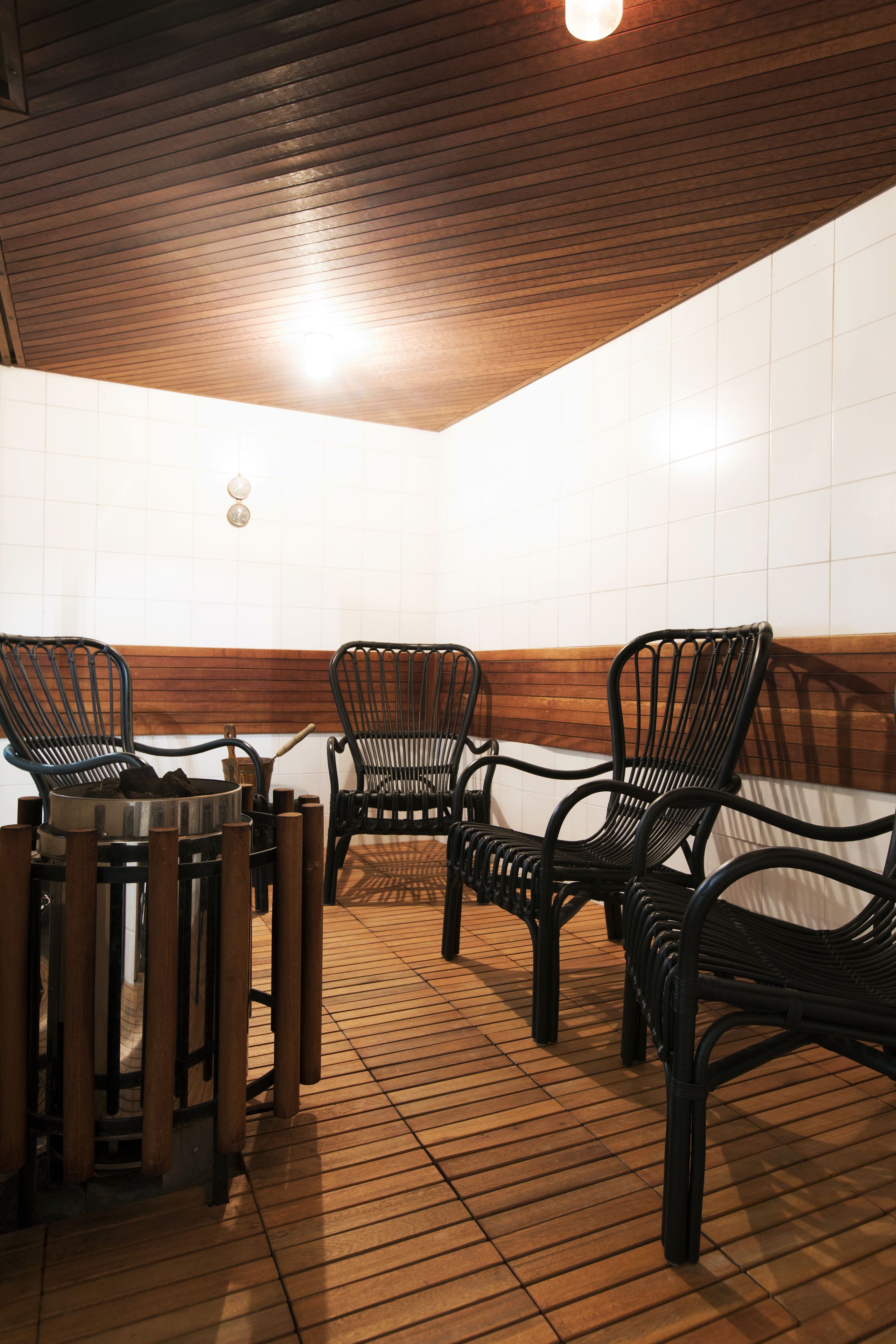A room with wooden flooring, a wooden ceiling, and a white tiled wall. There are four black chairs and a small table with a metal bowl.