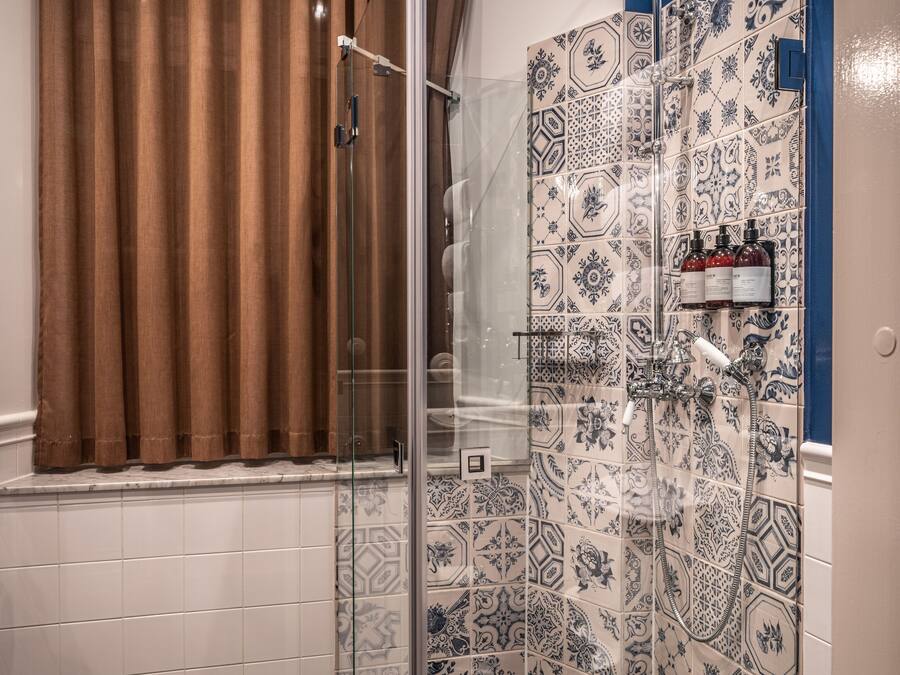 Un área de ducha con puerta de vidrio, azulejos estampados y una ventana con cortinas marrones.