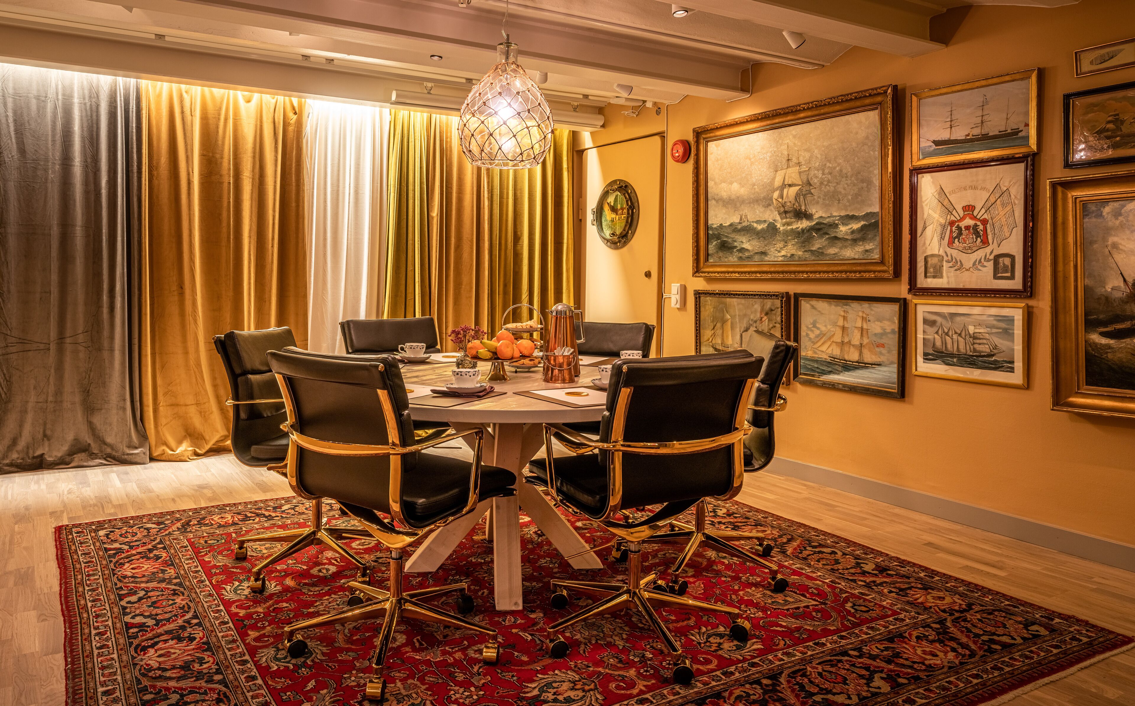 Een eetruimte met een ronde tafel, zwarte stoelen, een vloerkleed met een rood patroon, en maritieme kunstwerken aan de muren.