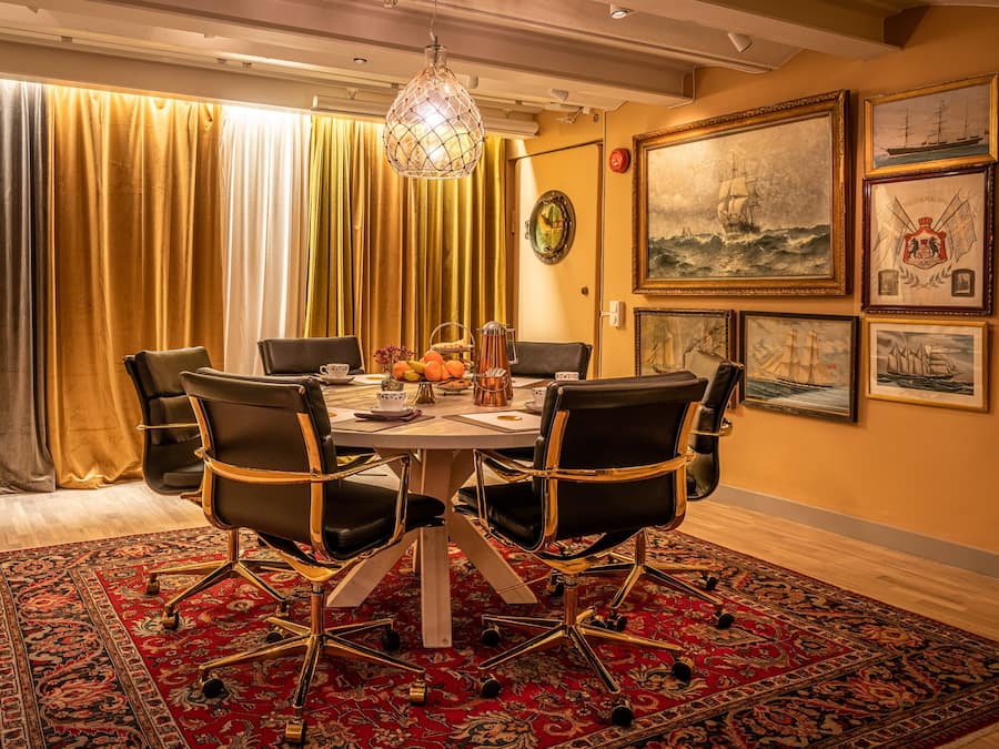 A dining area with a round table, black chairs, a red patterned rug, and maritime-themed artwork on the walls.