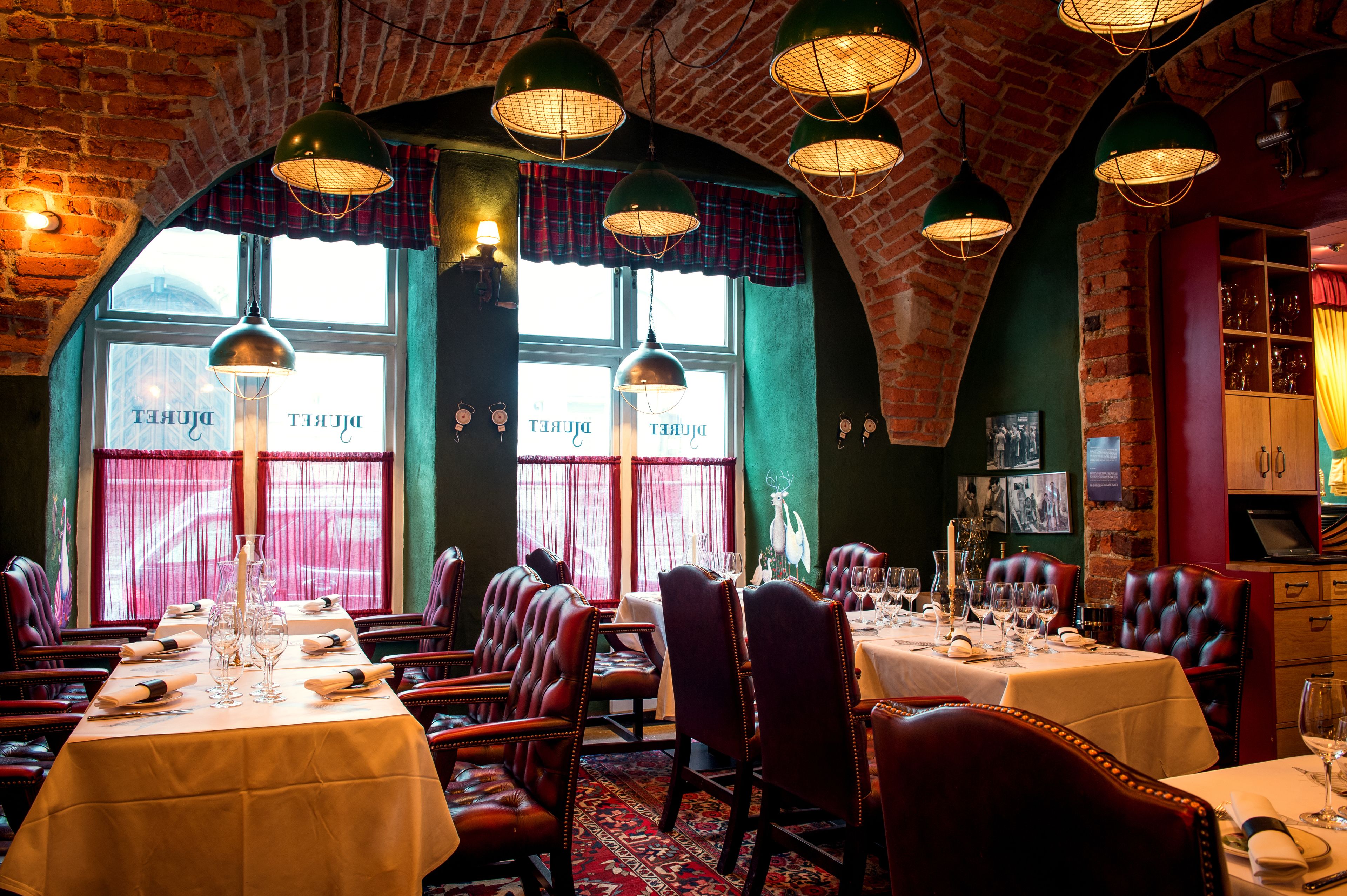Un restaurante con arcos de ladrillo, cortinas verdes y sillones de cuero. Las mesas están preparadas con manteles blancos y copas de vino.