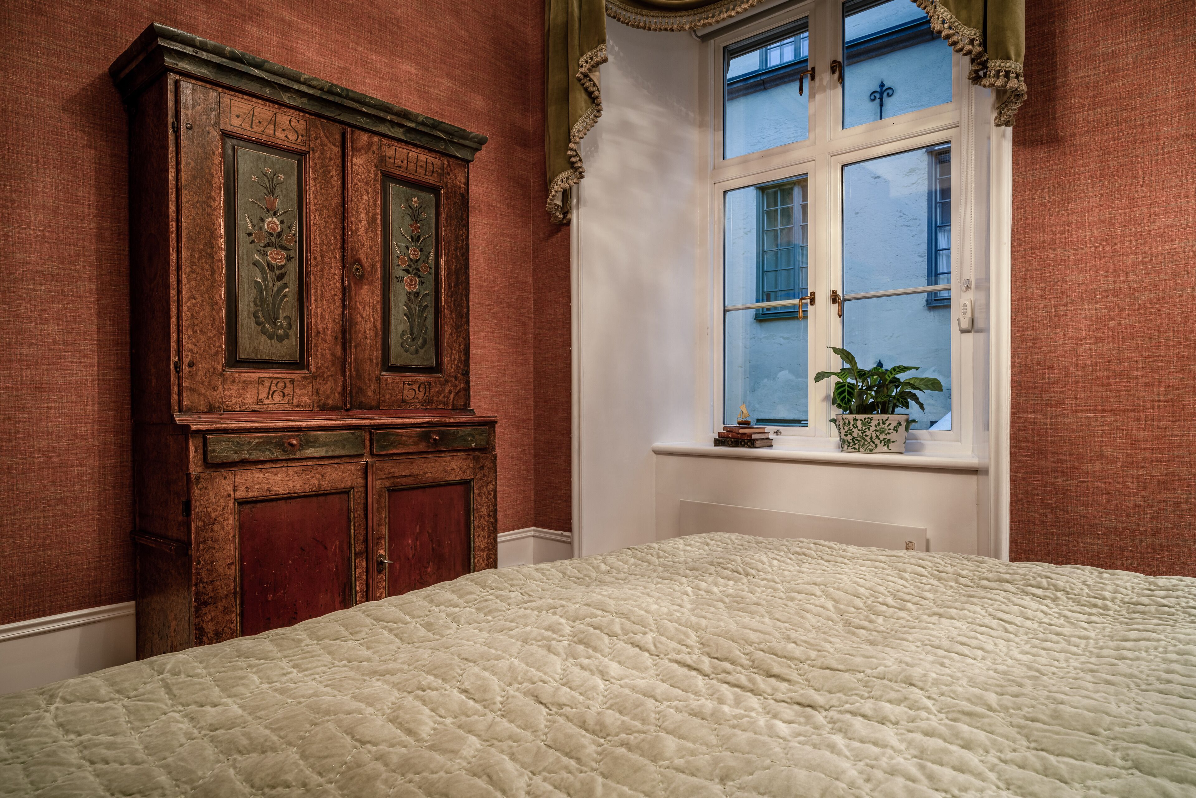 Une chambre avec un grand lit, une armoire en bois et une fenêtre agrémentée d’une plante.