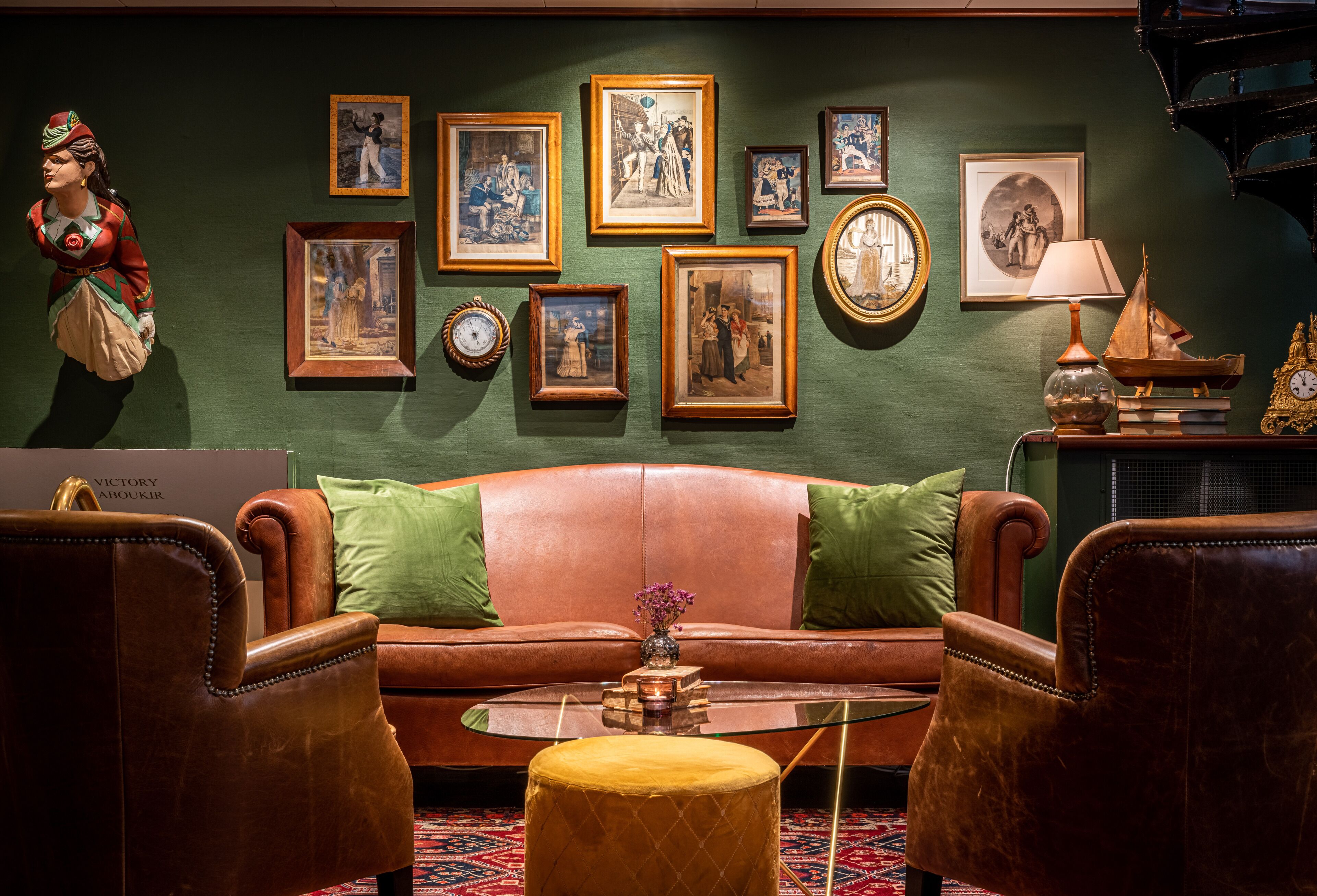 Ein Zimmer mit einer braunen Ledersofa, zwei grünen Kissen, einem Glastisch und einem kleinen, runden Hocker. An der Wand befinden sich gerahmte Bilder und links eine Statue.