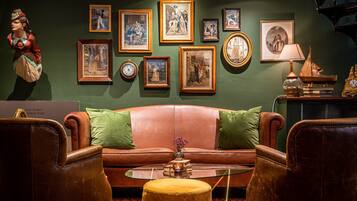 Ein Zimmer mit einer braunen Ledersofa, zwei grünen Kissen, einem Glastisch und einem kleinen, runden Hocker. An der Wand befinden sich gerahmte Bilder und links eine Statue.
