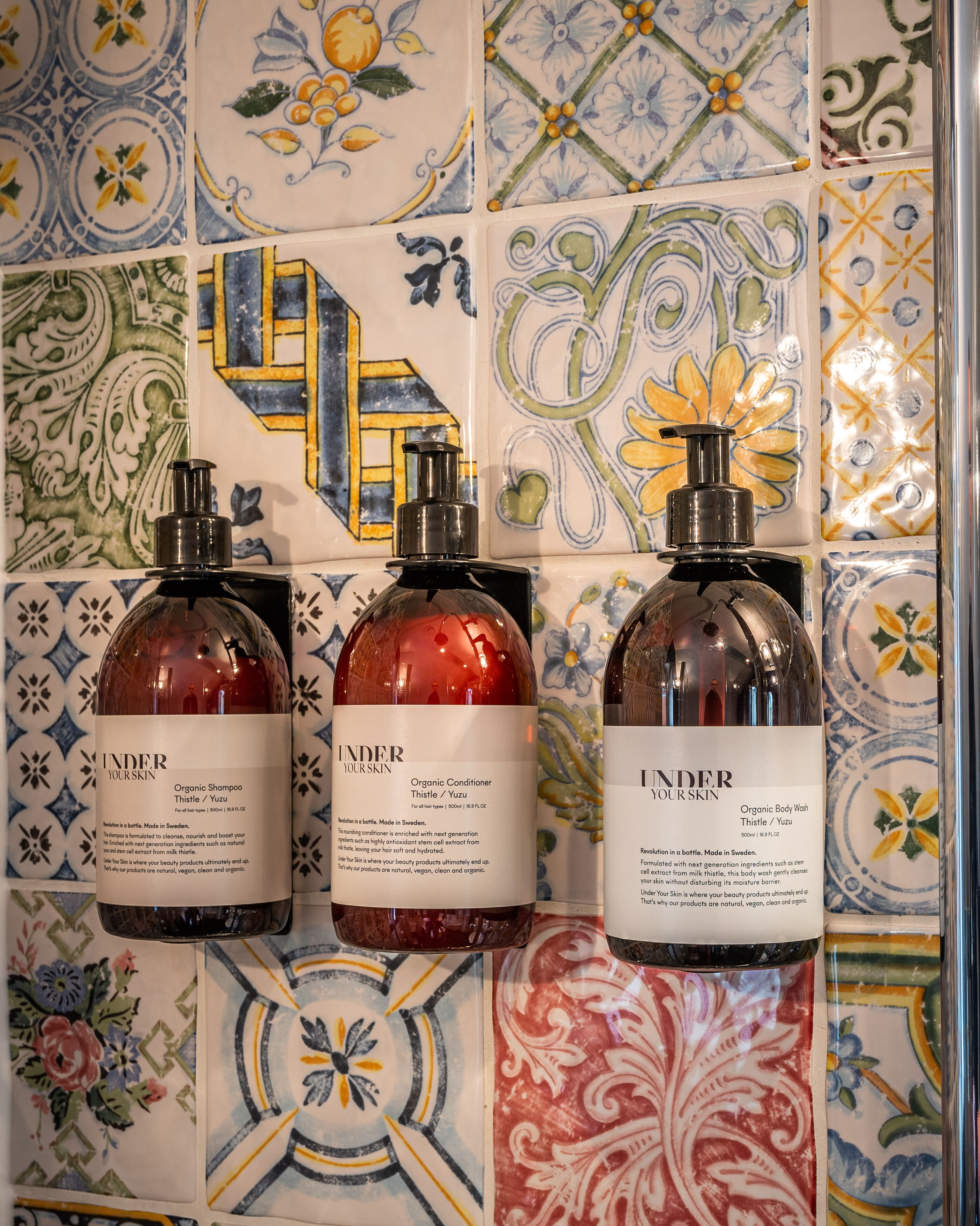 Tres botellas de productos de cuidado personal orgánico frente a una pared de azulejos con diversos diseños.