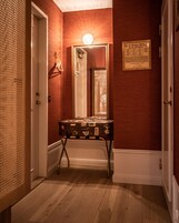 Un pasillo con piso de madera, una maleta antigua sobre un atril metálico, un espejo y una lámpara fijada a la pared.