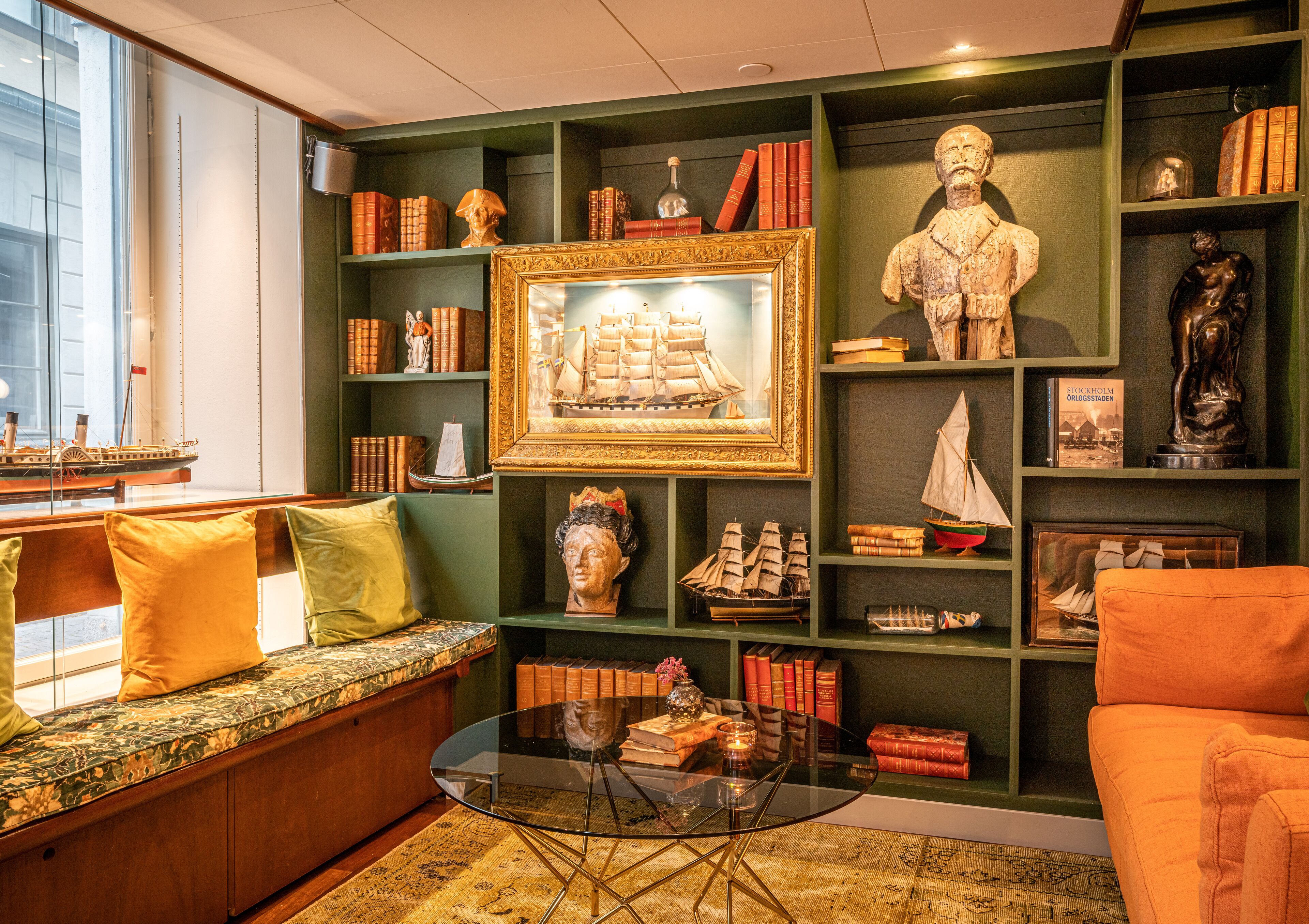 Un rincón acogedor para leer con un sofá, una mesa de centro de cristal y una estantería llena de libros y objetos decorativos.