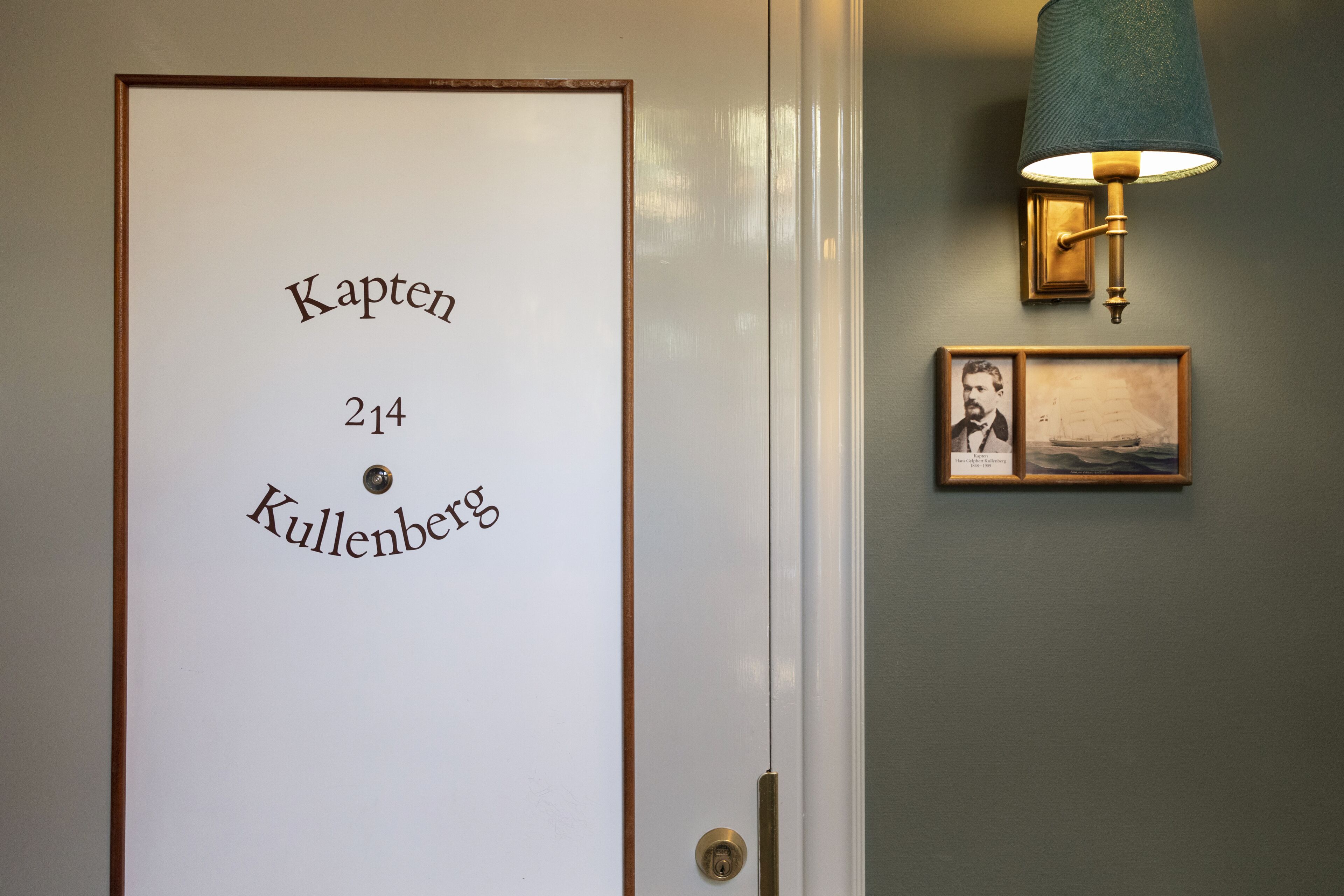 A door with the text "Kapten 214 Kullenberg" and a wall-mounted lamp with two framed pictures.