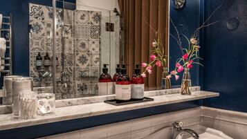A bathroom with a large mirror, two sinks, and a shower. There are toiletries on the counter and flowers in a vase.