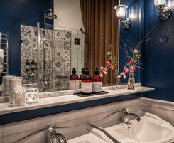 A bathroom with a large mirror, two sinks, and a shower. There are toiletries on the counter and flowers in a vase.