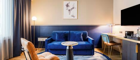 A modern hotel room with a blue sofa, a round coffee table, a brown leather chair, a small side table, a flat-screen TV, and a kitchen area with a coffee machine.