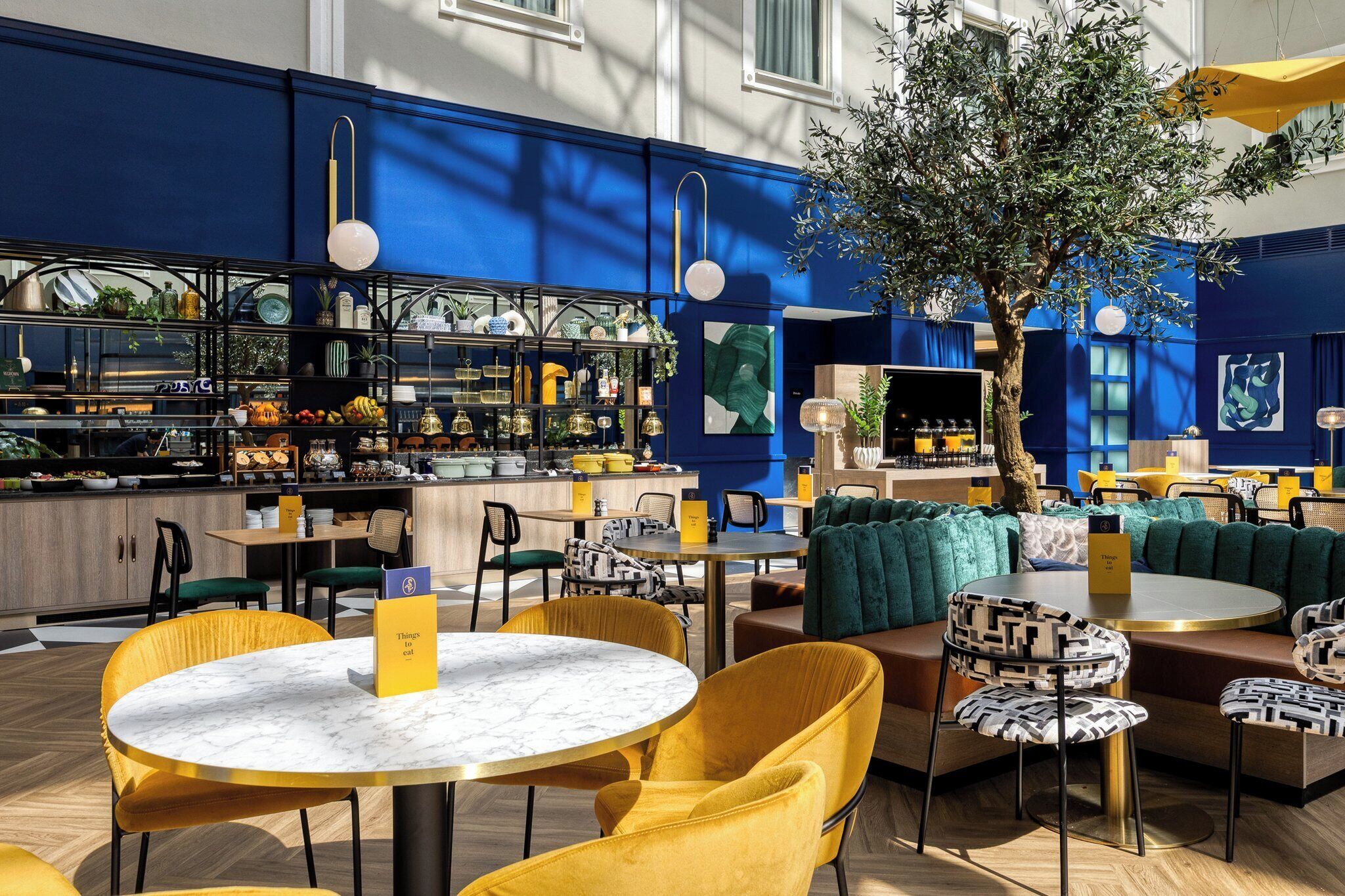 Un interior de restaurante moderno con mesas redondas, sillas amarillas y una zona de bar con estantes de utensilios de cocina.