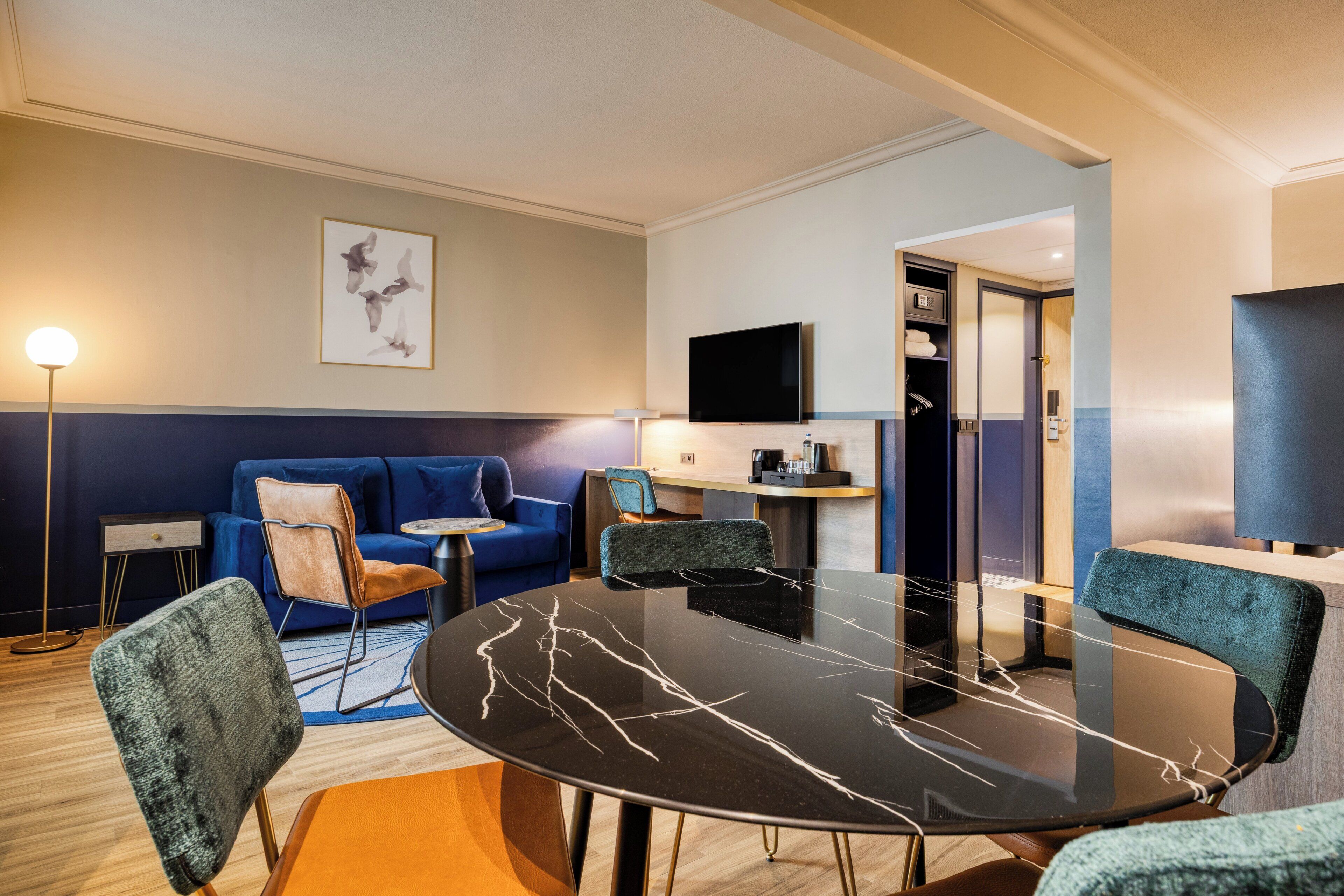 Una habitació d'hotel moderna amb una taula rodona de marbre negre, seients amb tapisseria blava i marró, un televisor de pantalla plana i una obra d'art emmarcada a la paret.