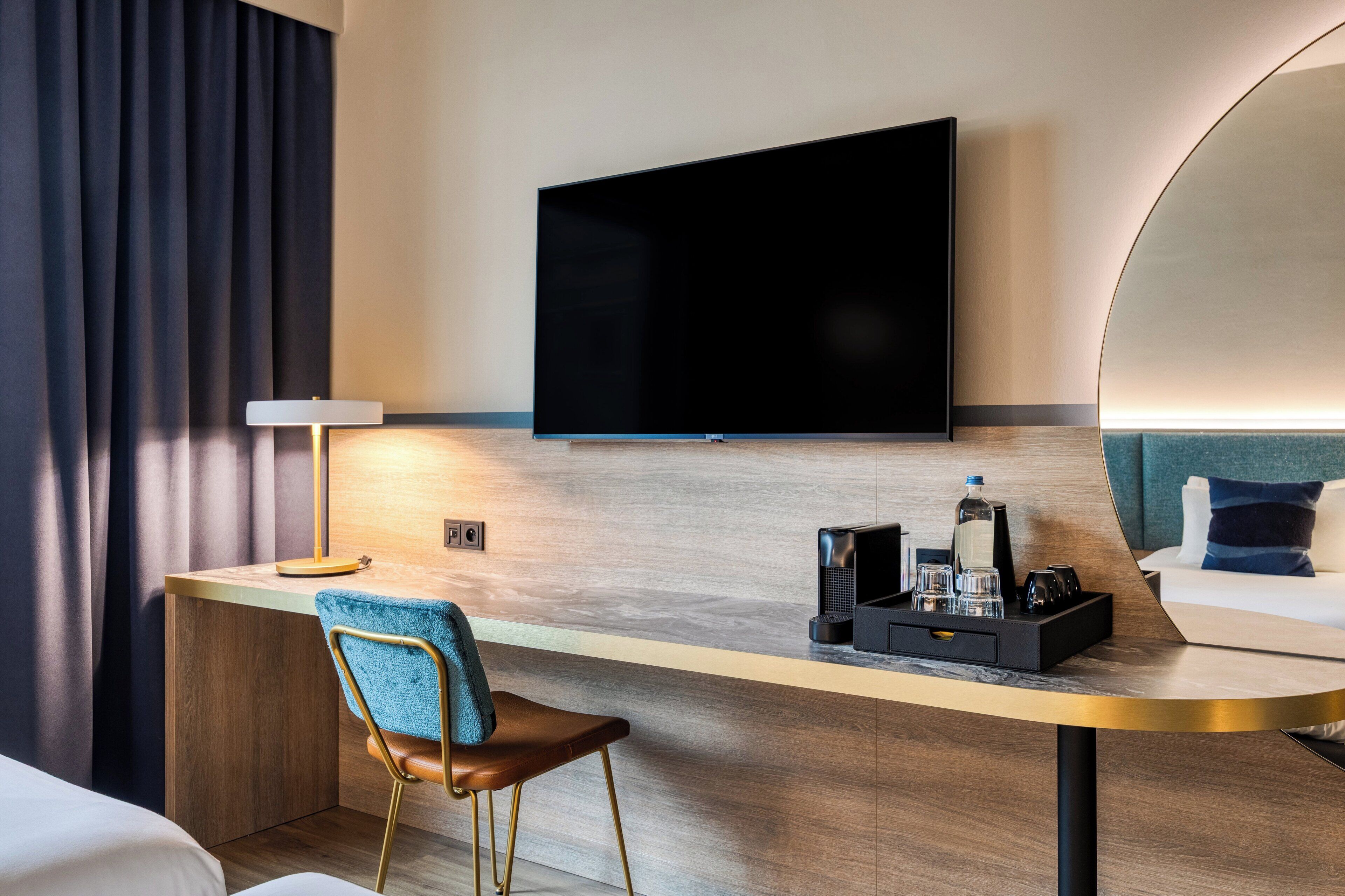 A hotel room with a flat-screen TV mounted on the wall, a desk with a chair, a bedside table with a lamp, and a bed with a headboard.