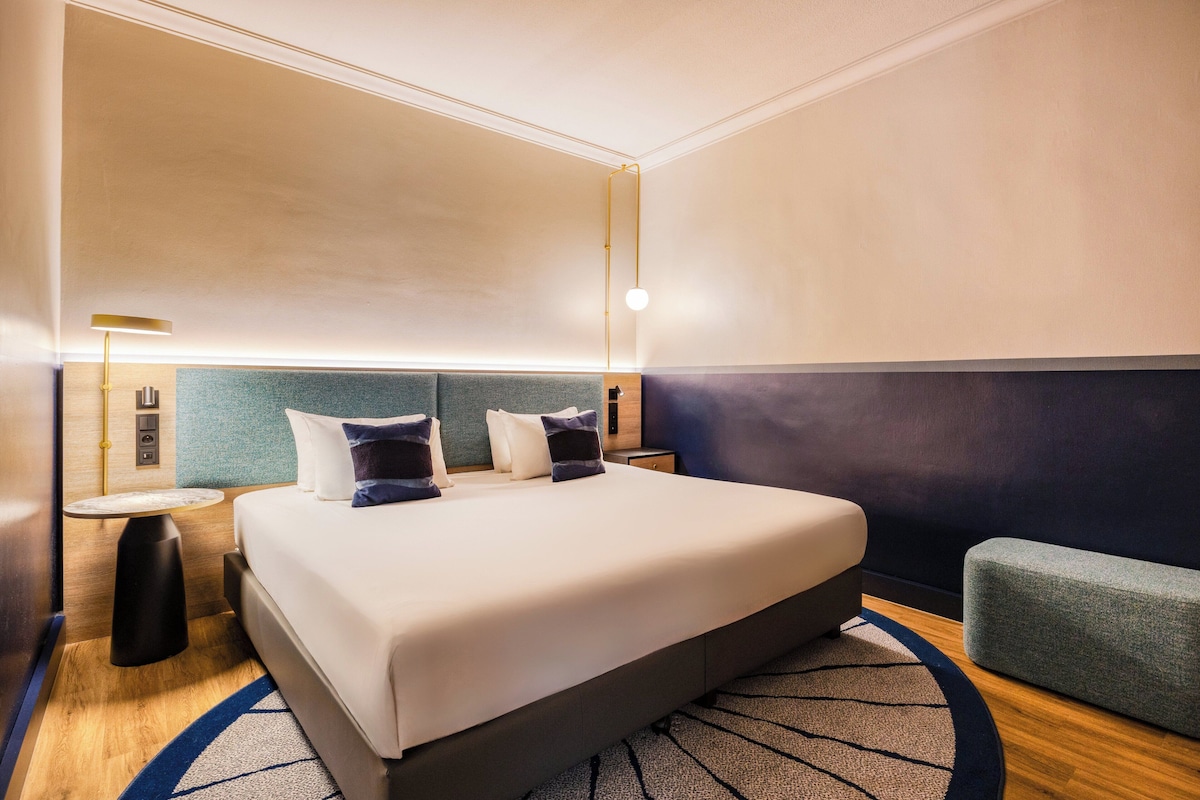A hotel room with a large bed, bedside tables, a round rug, and a seating area.