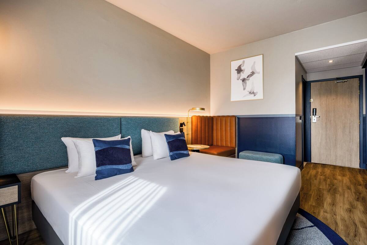 A hotel room with a large bed, a desk, a chair, and a door.