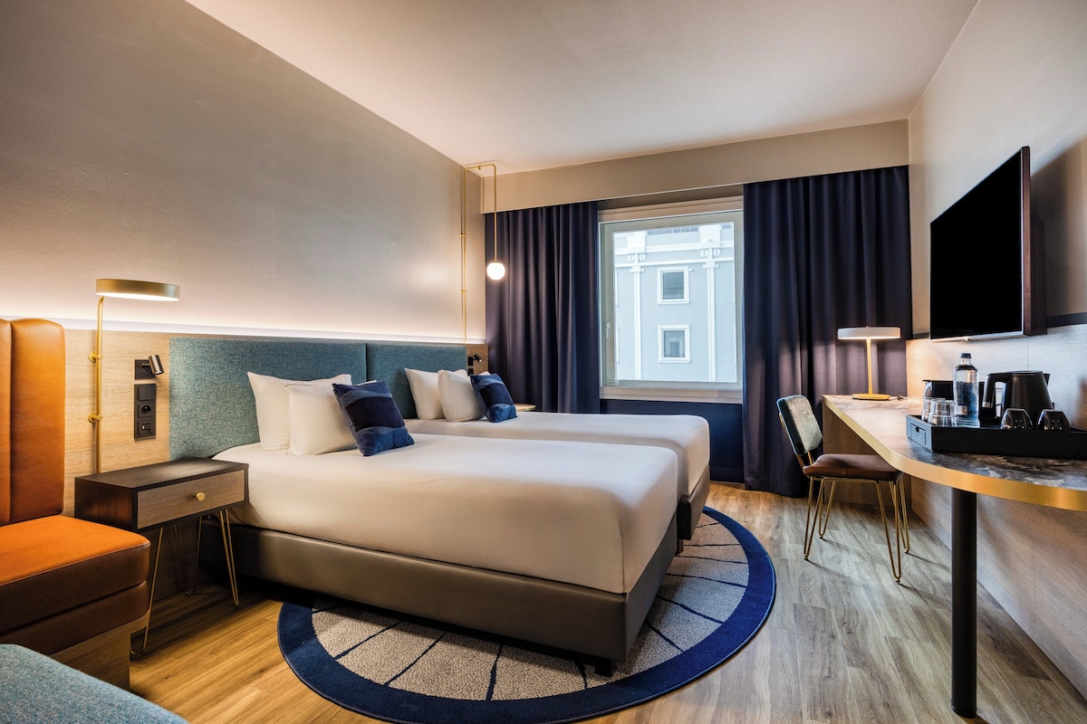 A hotel room with a large bed, a desk, a chair, a TV, and a window with curtains.