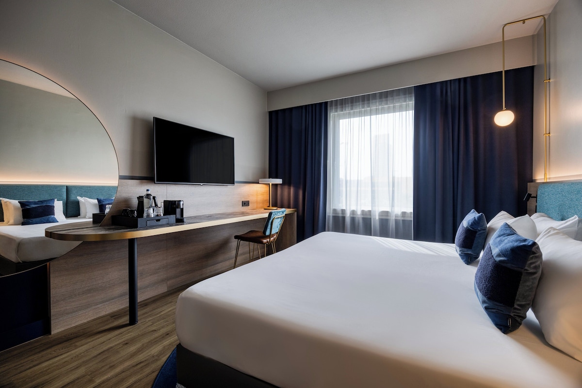 A hotel room with a bed, a desk, a chair, a TV, and a window with curtains.
