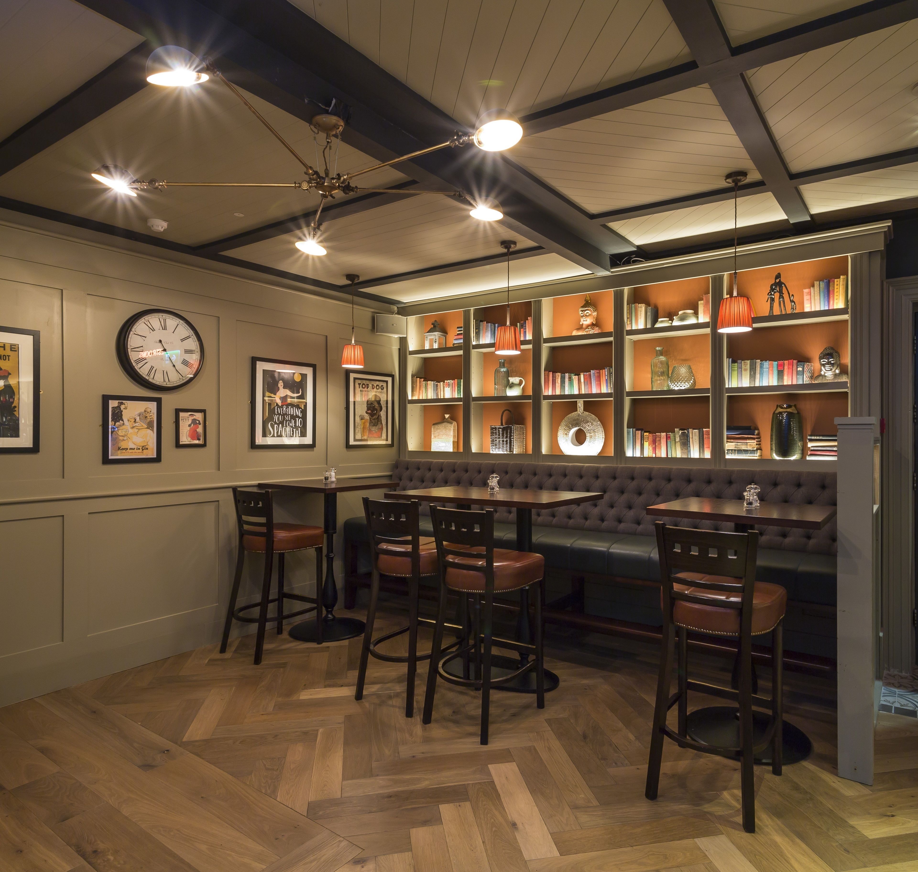 A cozy bar area with wooden flooring, a round table, and bar stools. There are bookshelves filled with books and decorative items, and a clock on the wall.