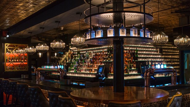 Zona de bar con diversas opciones de asientos, una estantería bien surtida de licores y una lámpara decorativa central.