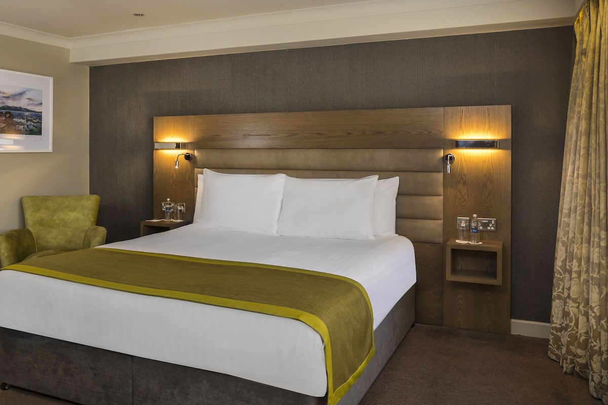 Ein Hotelzimmer mit einem großen Bett, einem grünen Sessel, einem Kopfteil mit integrierter Beleuchtung und einem gerahmten Bild an der Wand.