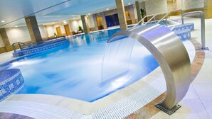 Una piscina interior con borde curvo, suelo de baldosas y una zona de estar con sillas y una mesa.