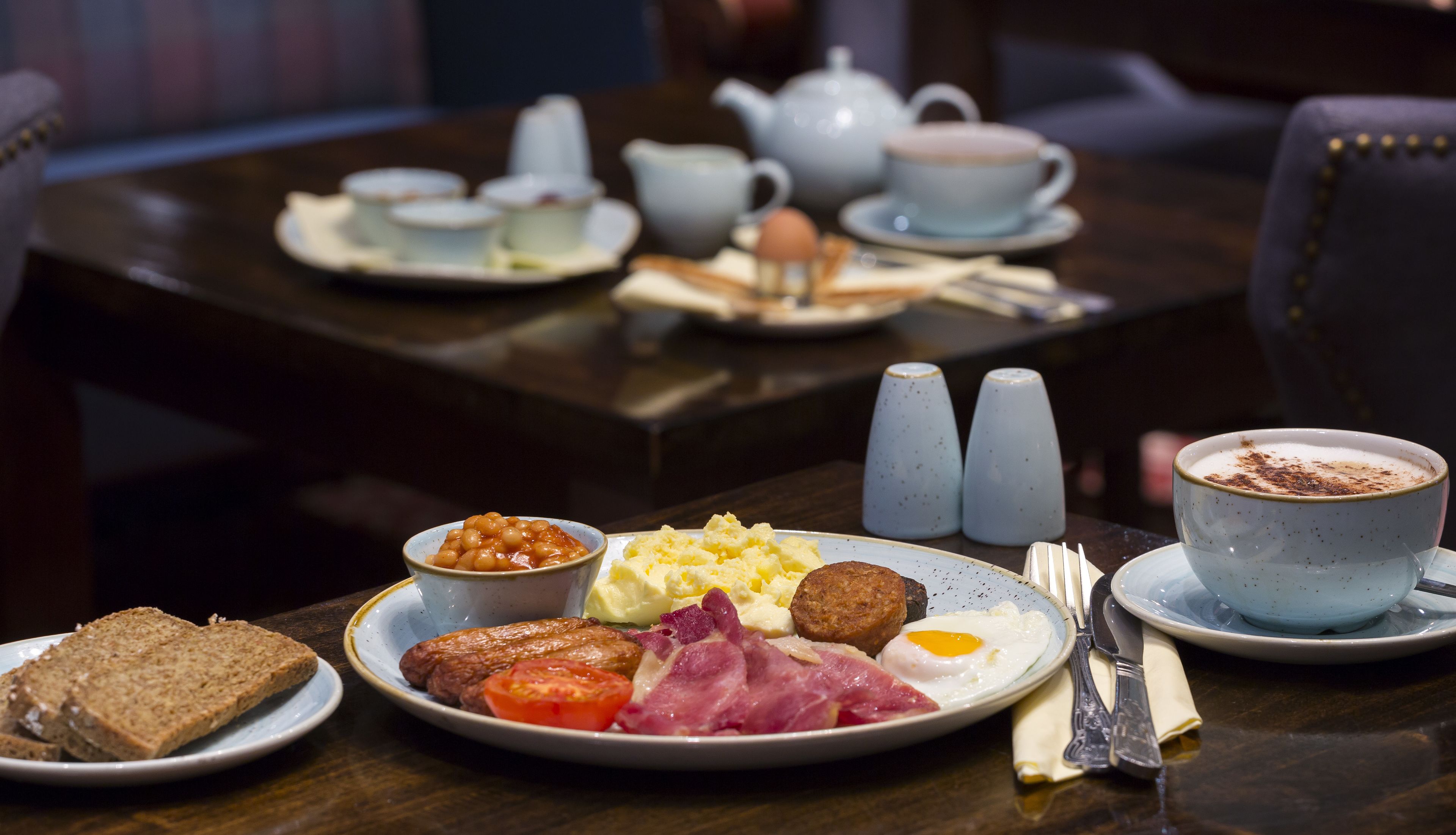 Un desayuno con platos de huevos, tocino, tomates y tostadas, junto con una taza de café y una tetera sobre una mesa.