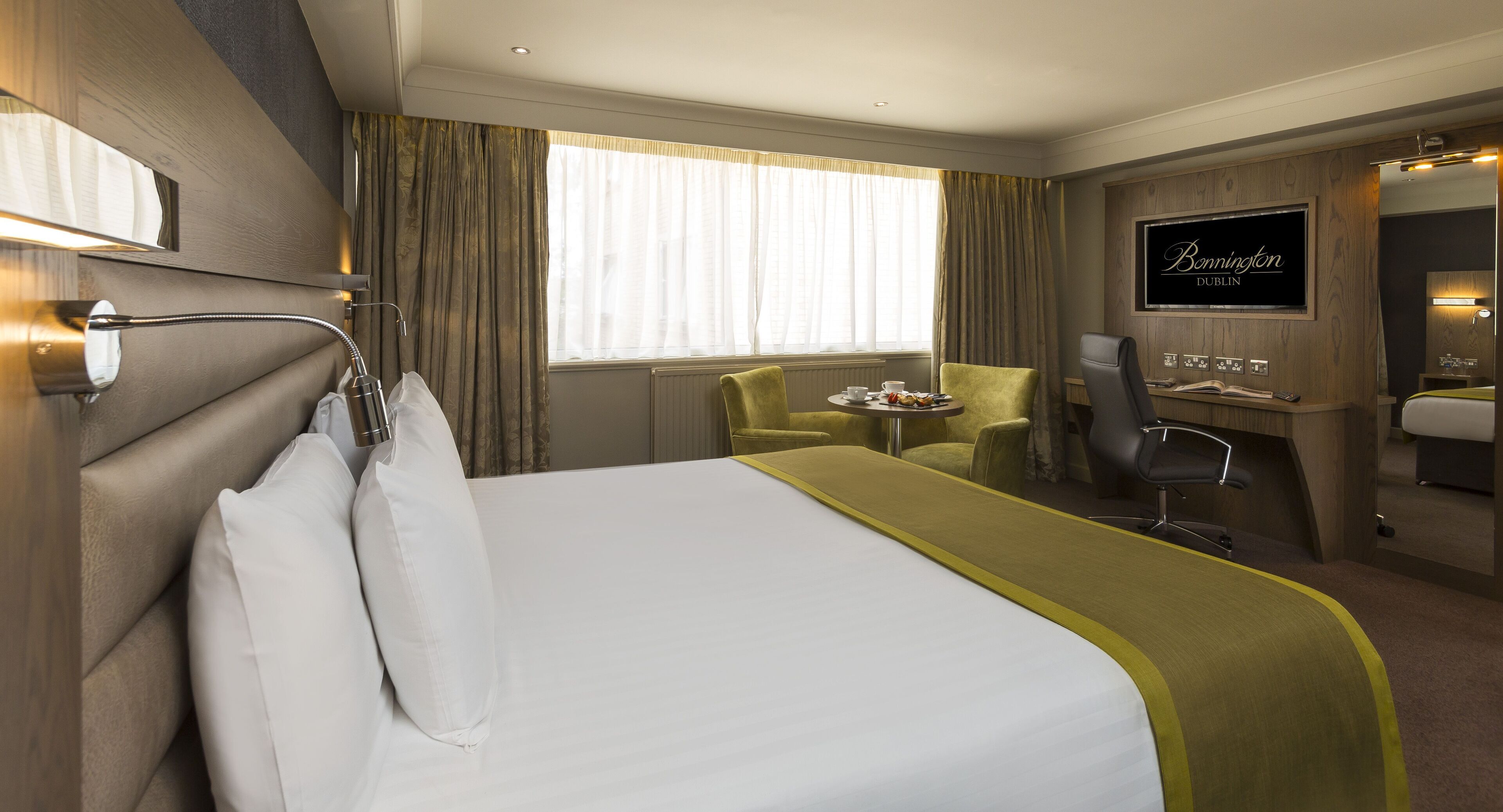 Quarto de hotel com cama, escrivaninha, cadeira, televisão e janela com cortinas.