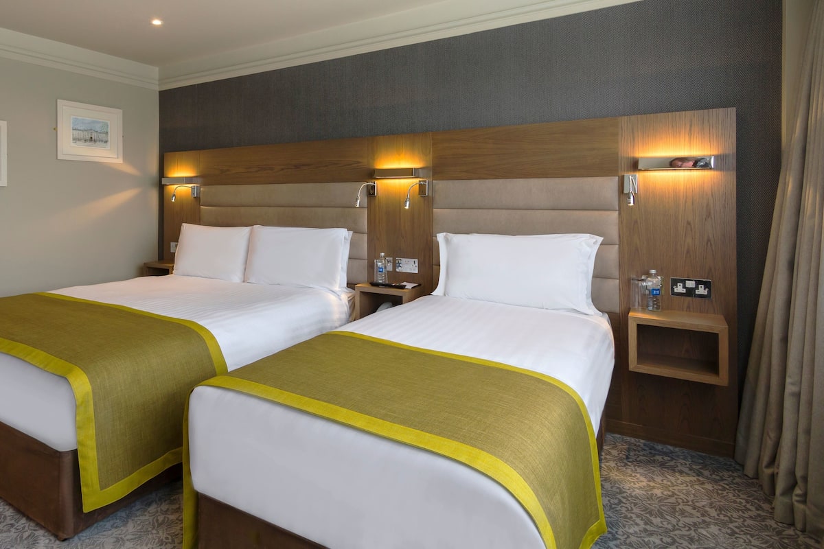 Ein Hotelzimmer mit zwei Betten, jeweils mit einem Holz-Kopfteil und einem Nachttisch. An den Wänden hängen Bilder in Rahmen, und eine kleine Ablagefläche zeigt eine Flasche und ein Glas.