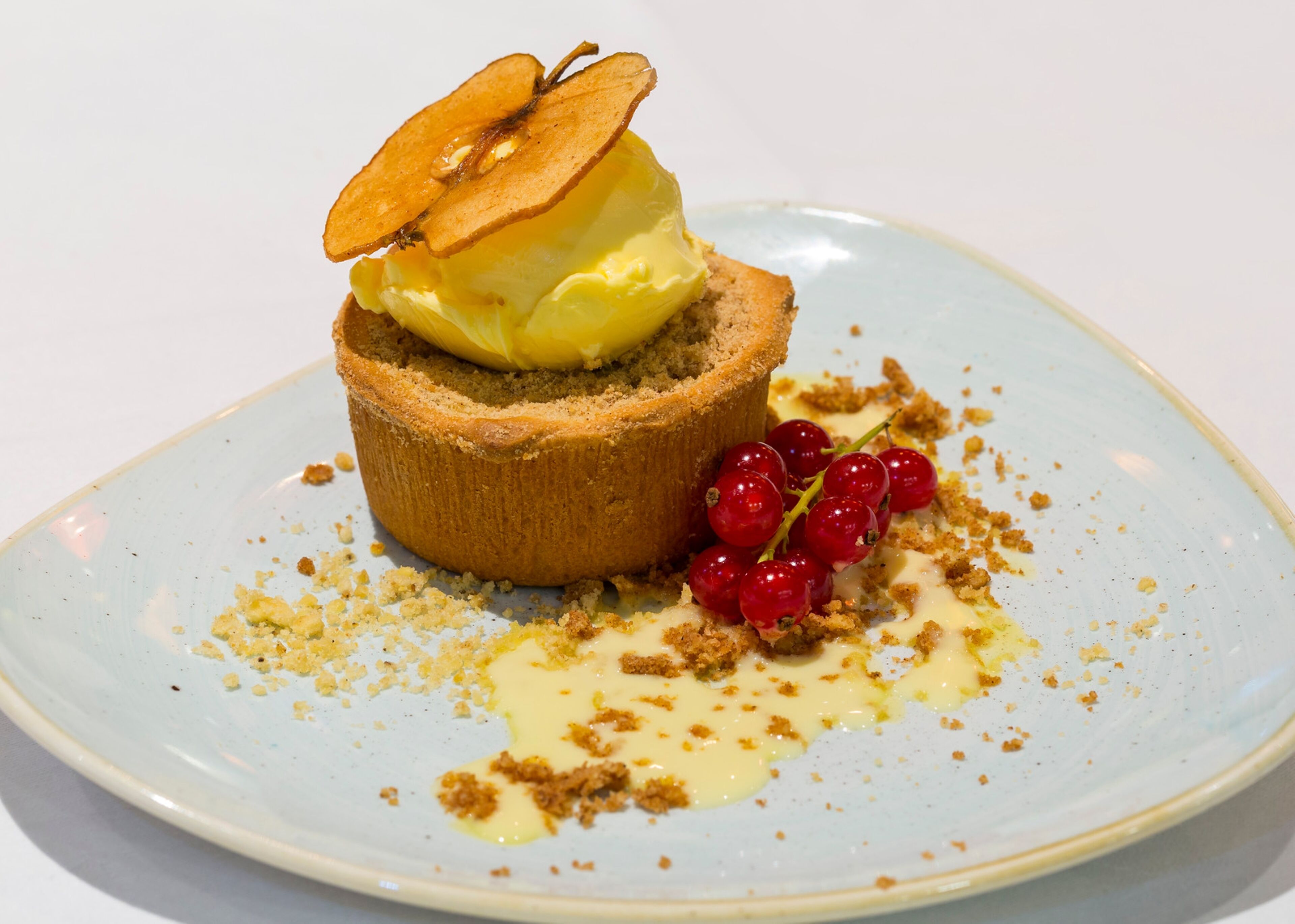 A dessert on a plate with a crumbly base, topped with a slice of cake and a scoop of yellow ice cream, accompanied by red currants and a drizzle of sauce.