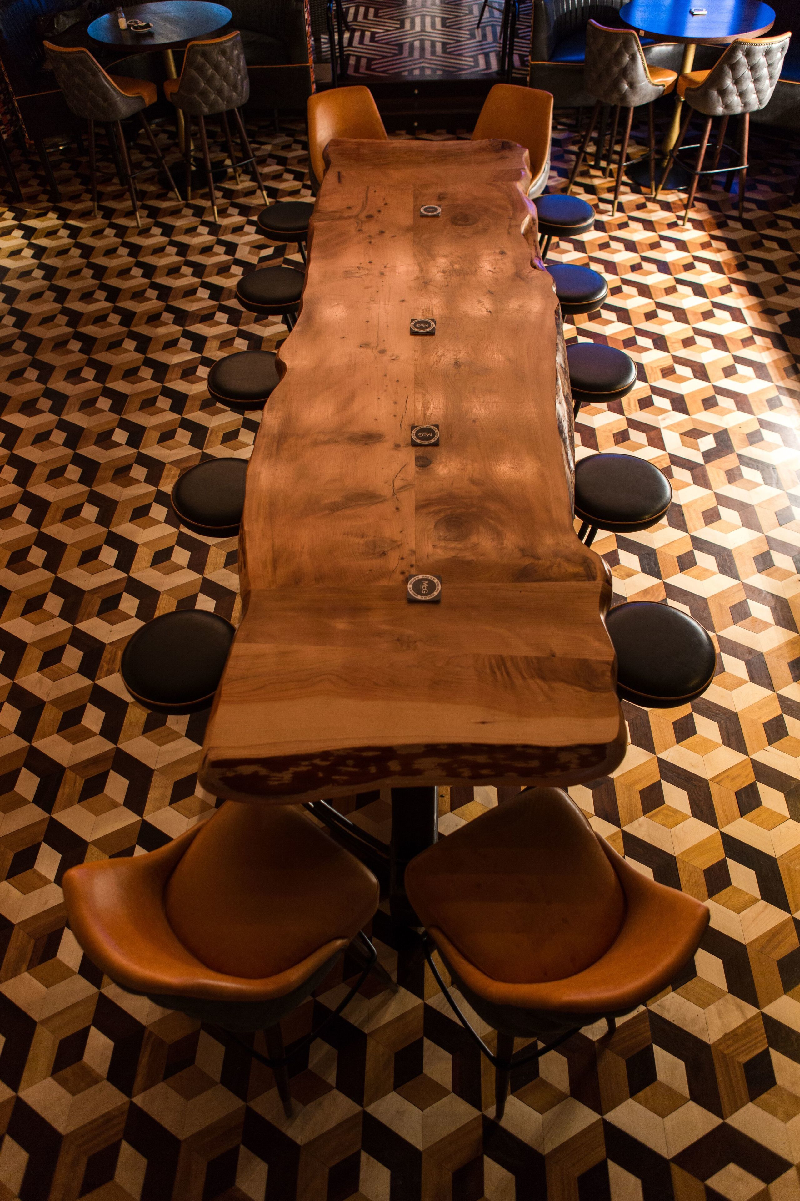 Une longue table en bois, entourée de six chaises noires à coussins, se trouve au-dessus d’un sol à motifs. On aperçoit également quelques tables et chaises à l’arrière-plan.