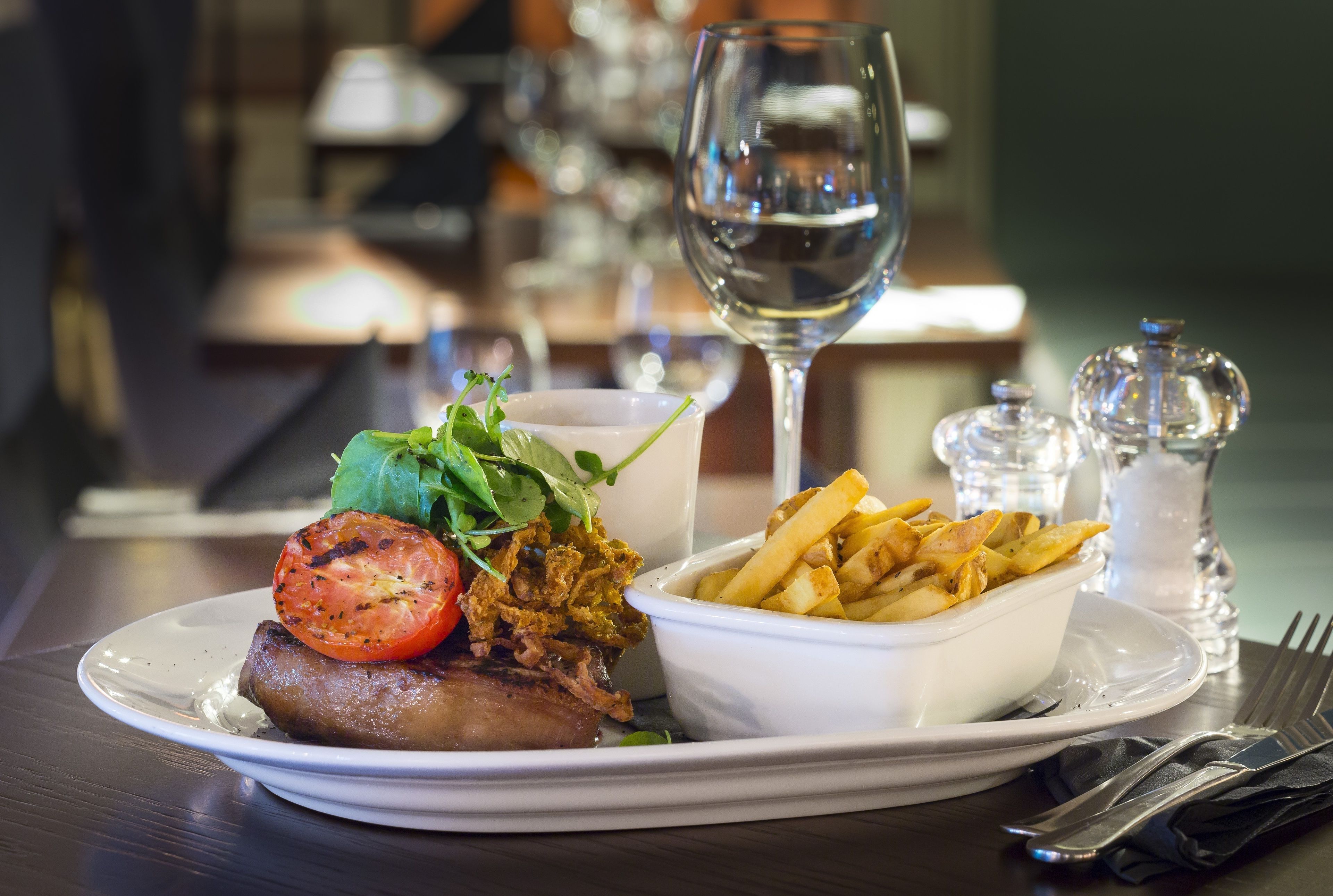 A plate of food with a steak, a tomato, and fries, a glass of water, and salt and pepper shakers on a table.