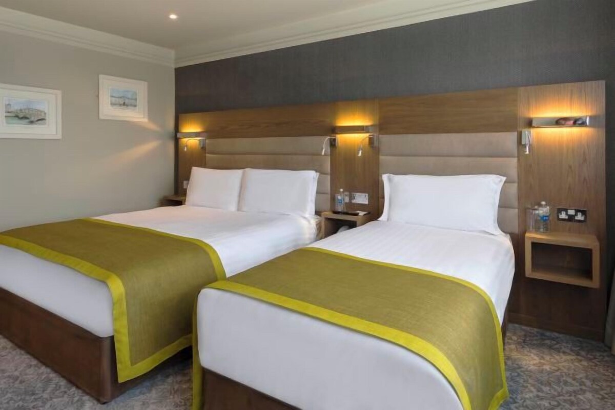 Habitación de hotel con dos camas, cabecero de madera, mesitas de noche y una pequeña mesita auxiliar.