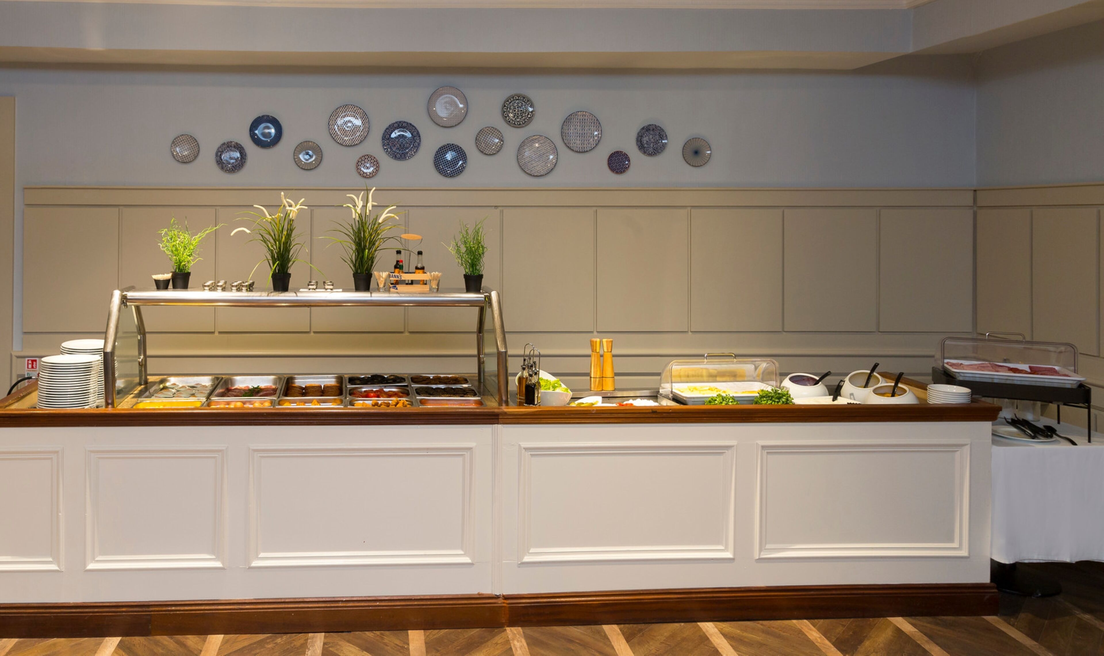 A buffet area with a variety of food options, a display of decorative plates on the wall, and potted plants.