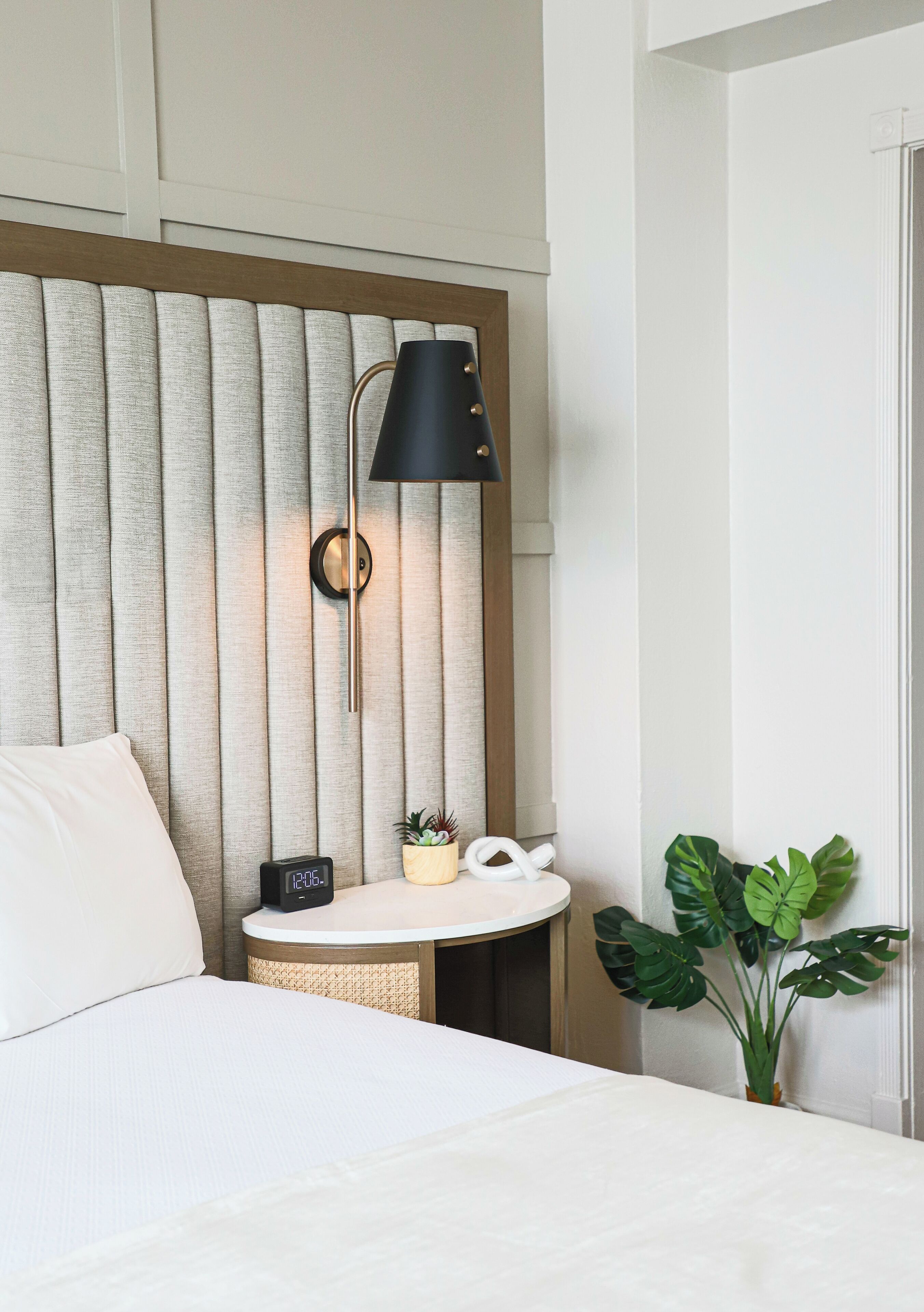 Une chambre d’hôtel avec un lit, une table de chevet avec une horloge, une lampe et une plante en pot.