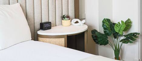 A hotel room with a bed, a bedside table with a clock, a lamp, and a potted plant.