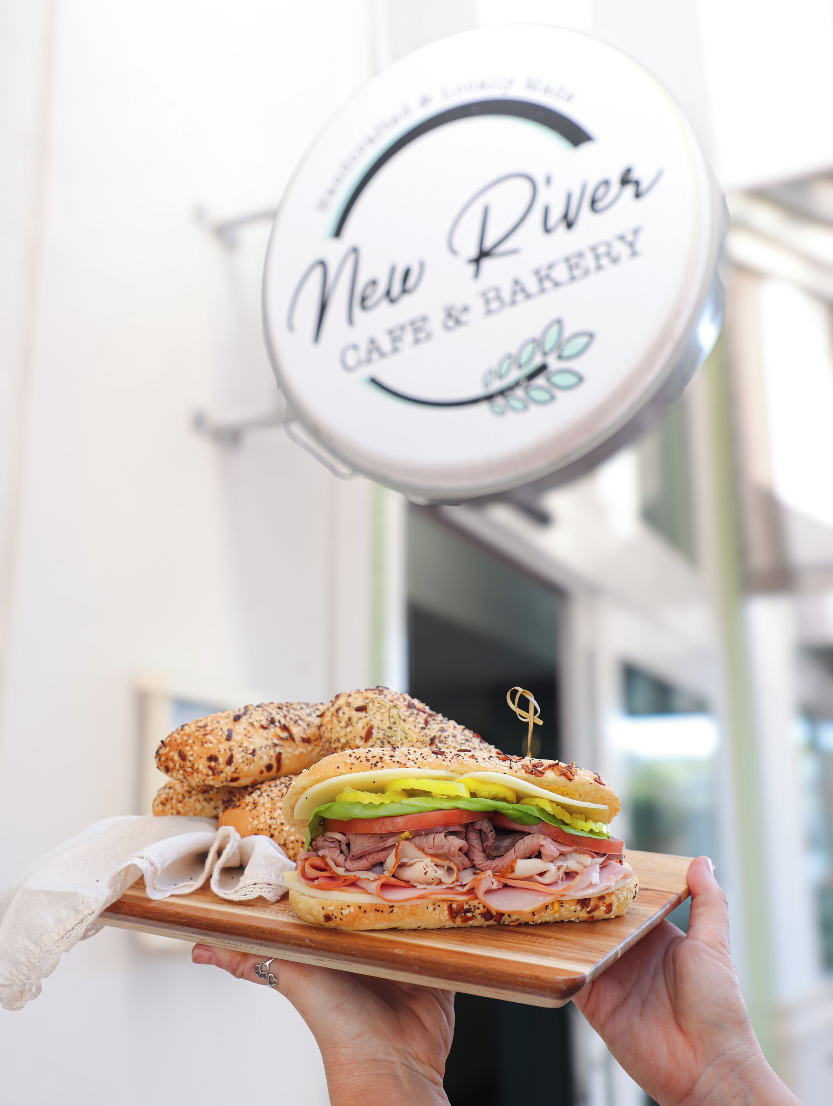 A sandwich on a wooden board held by a person, with a sign reading "New River Cafe & Bakery" in the background.