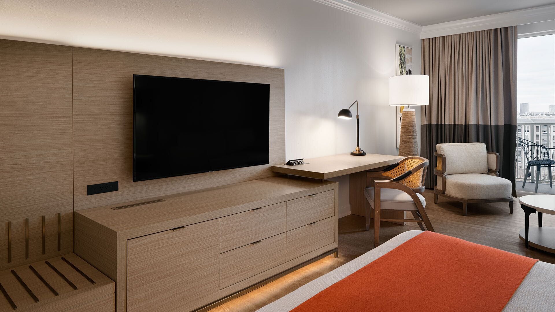Hotelkamer met flatscreen-tv, bureau, stoel, lamp en uitzicht op de stad.