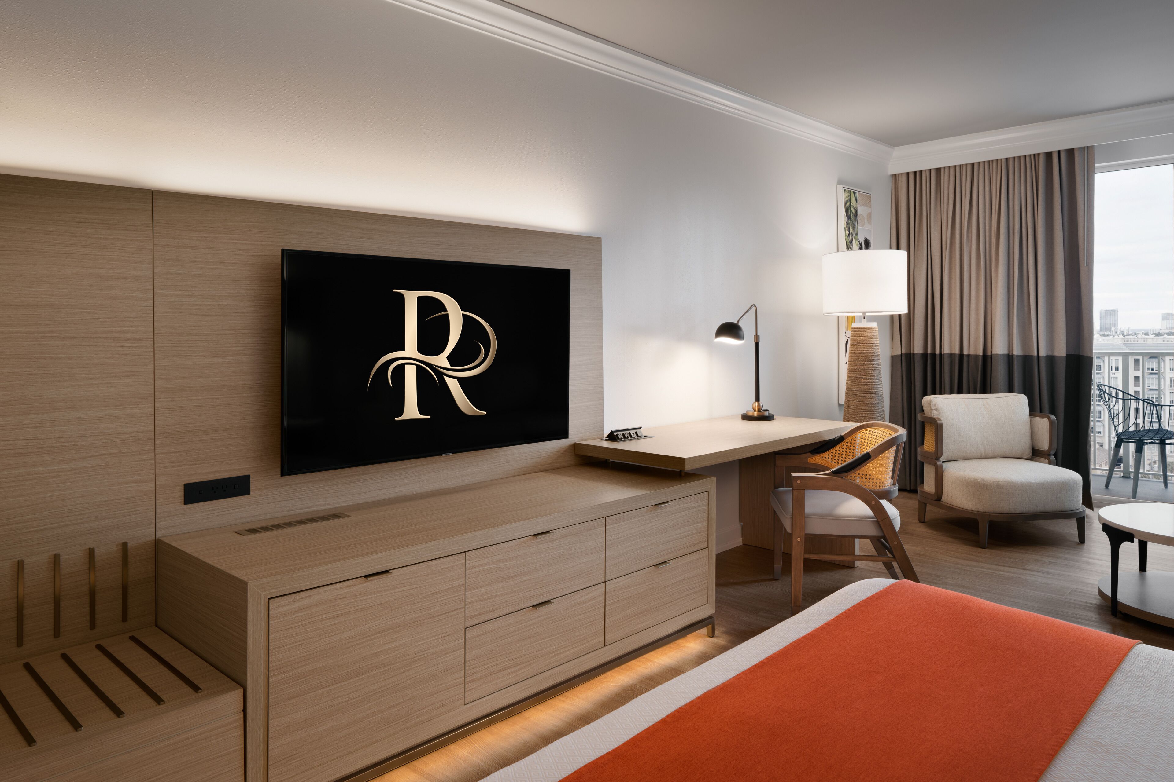 A modern hotel room with a large bed, a desk, two chairs, a TV mounted on the wall, and a view of the city through the window.