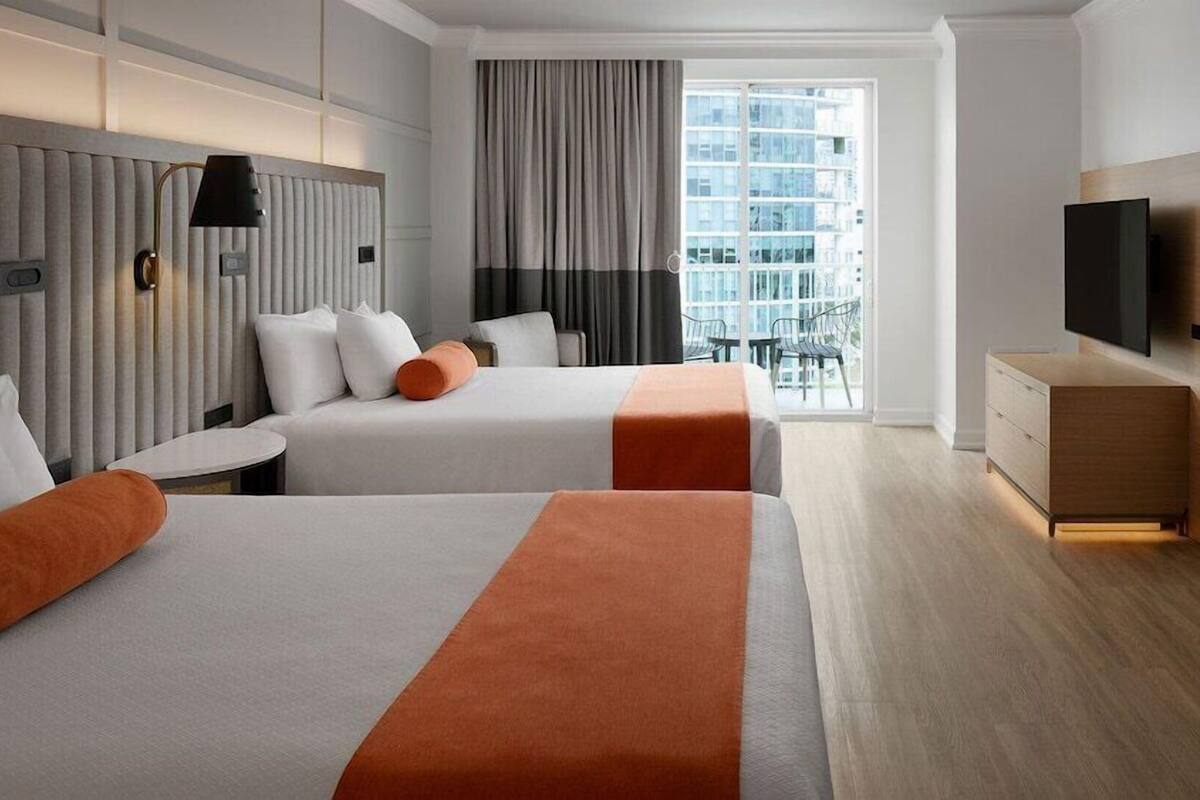 A hotel room with two beds, a TV, a desk, and a view of the city.