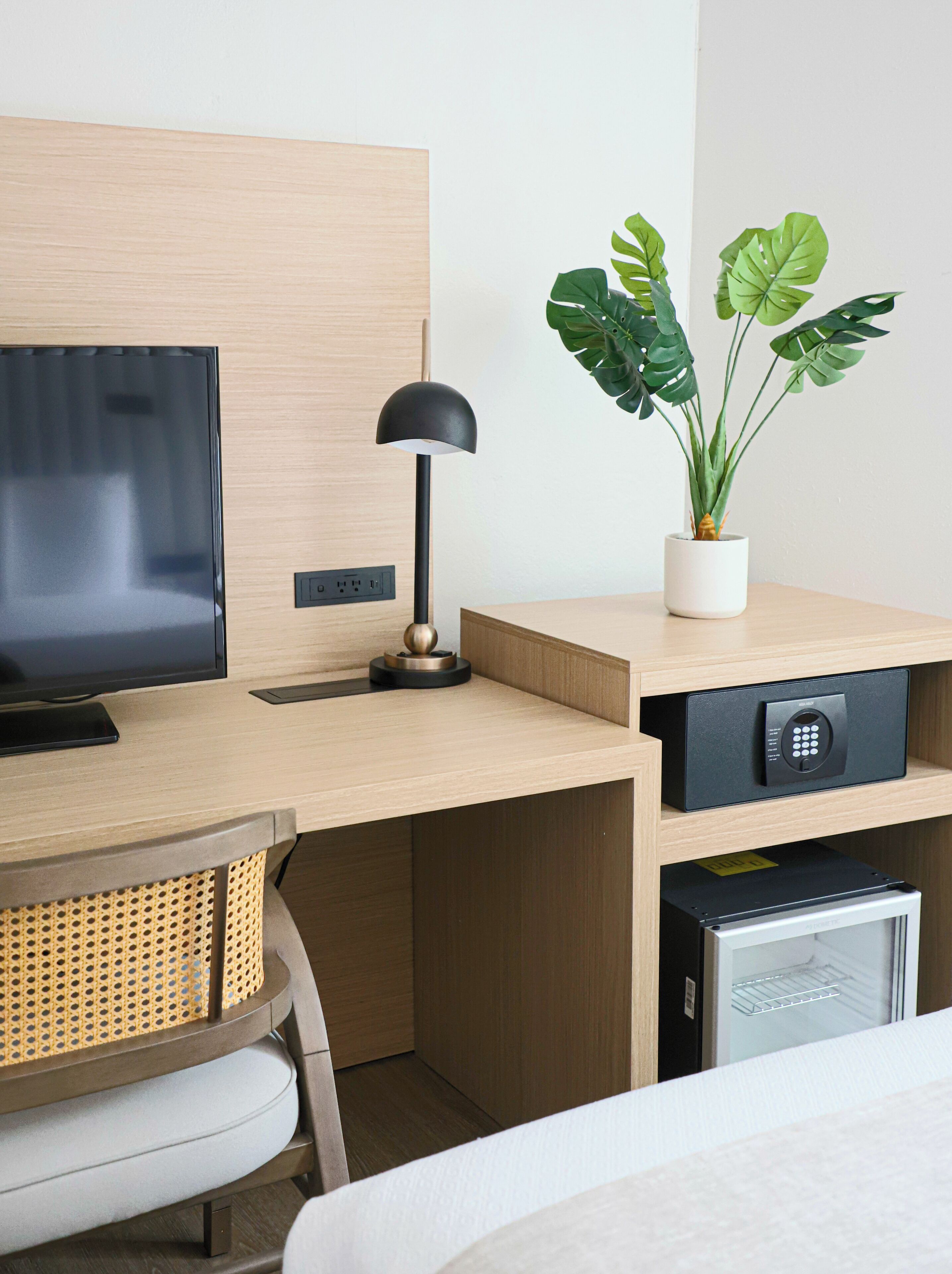 Une chambre d’hôtel moderne équipée d’un bureau en bois, d’un téléviseur à écran plat, d’une lampe, d’une plante en pot, d’un coffre-fort et d’un mini-bar.