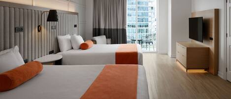 A hotel room with two beds, a TV, a desk, and a balcony with a view of the city.