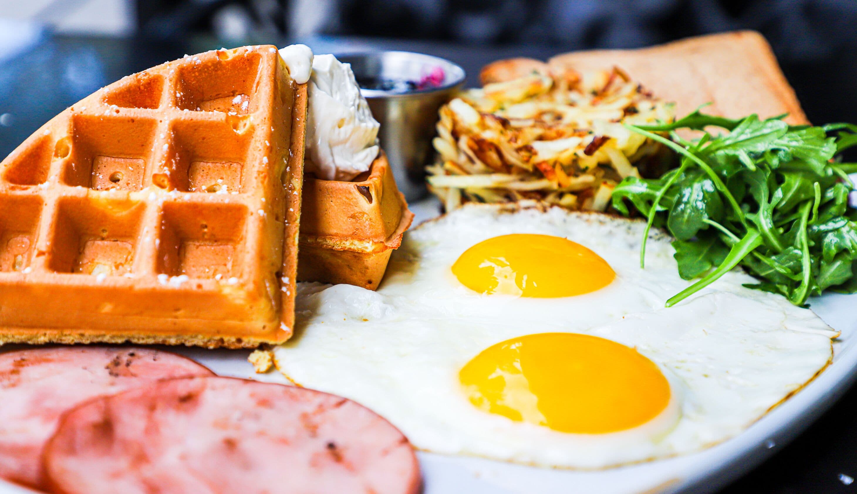 A plate with a waffle, two fried eggs, a slice of ham, and a side salad.