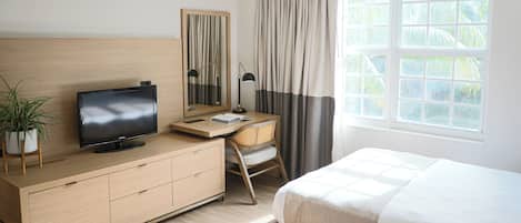 A hotel room with a bed, a desk with a TV, a chair, a dresser, a mirror, and a window with curtains.